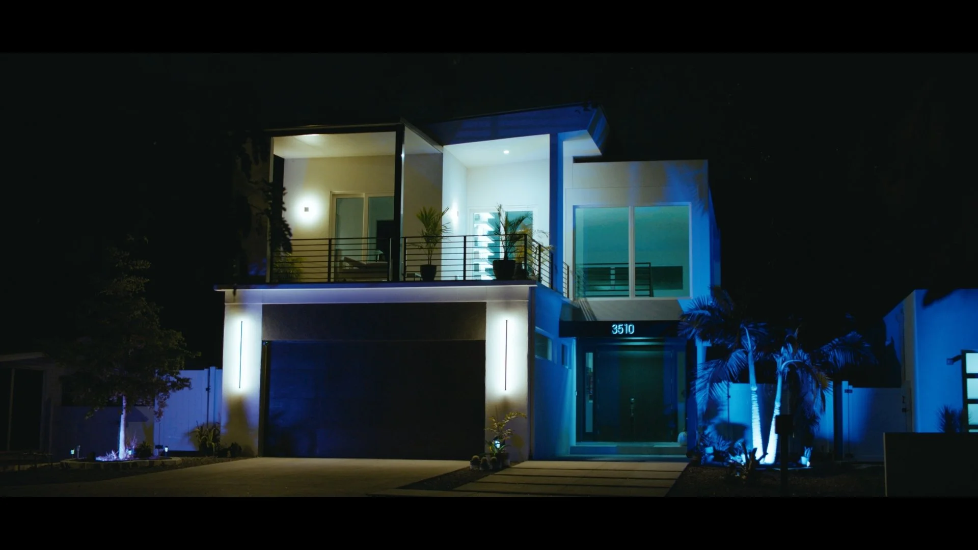 Modern two-story house at night with exterior lighting, plants on balcony, house number 3510, illuminated by blue and white lights.