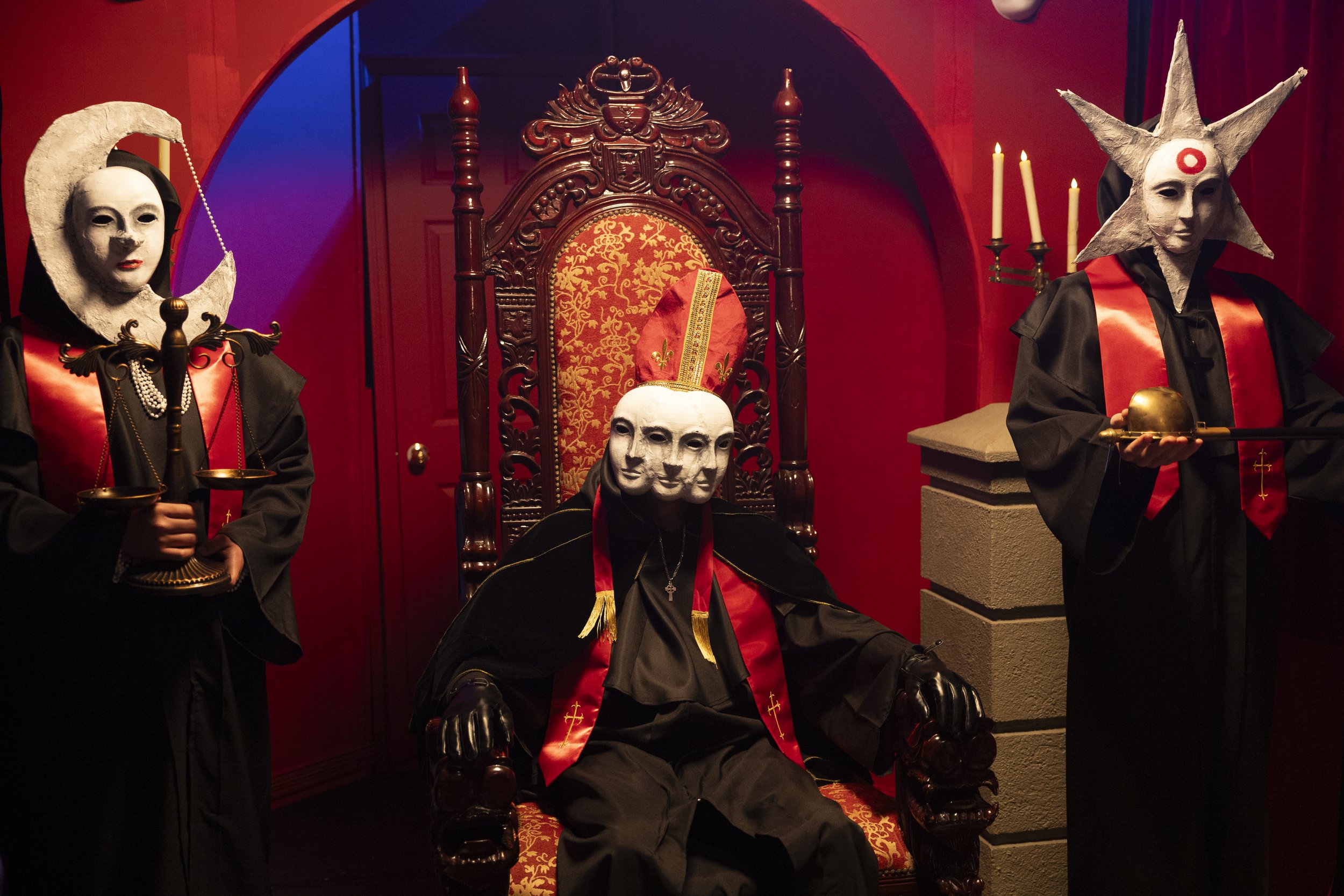 Three figures in theatrical costumes, one seated on an ornate wooden throne with a red and gold upholstered backrest, wearing a mask with multiple faces, and two standing figures in black robes with elaborate masks and star and moon motifs, holding ceremonial objects, in a red-walled room with candles.
