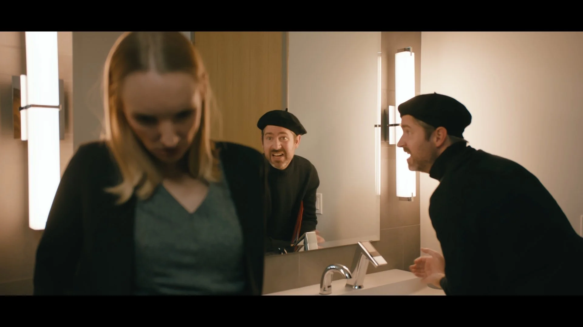 Three people are in a bathroom, two men wearing black berets are angry and shouting at a woman with blonde hair, who is looking down.