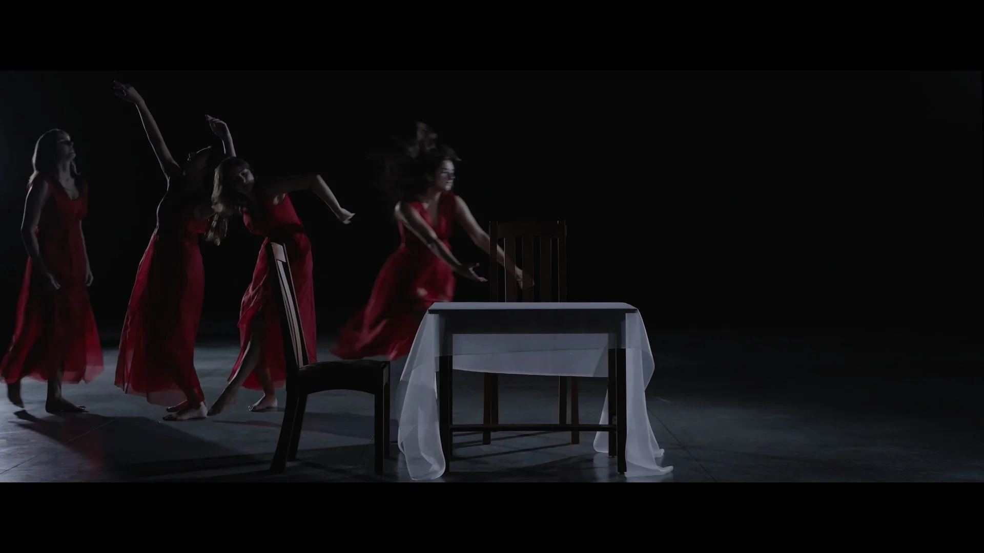 Five women in red dresses dancing around a table with a white tablecloth and four chairs in a dark room.