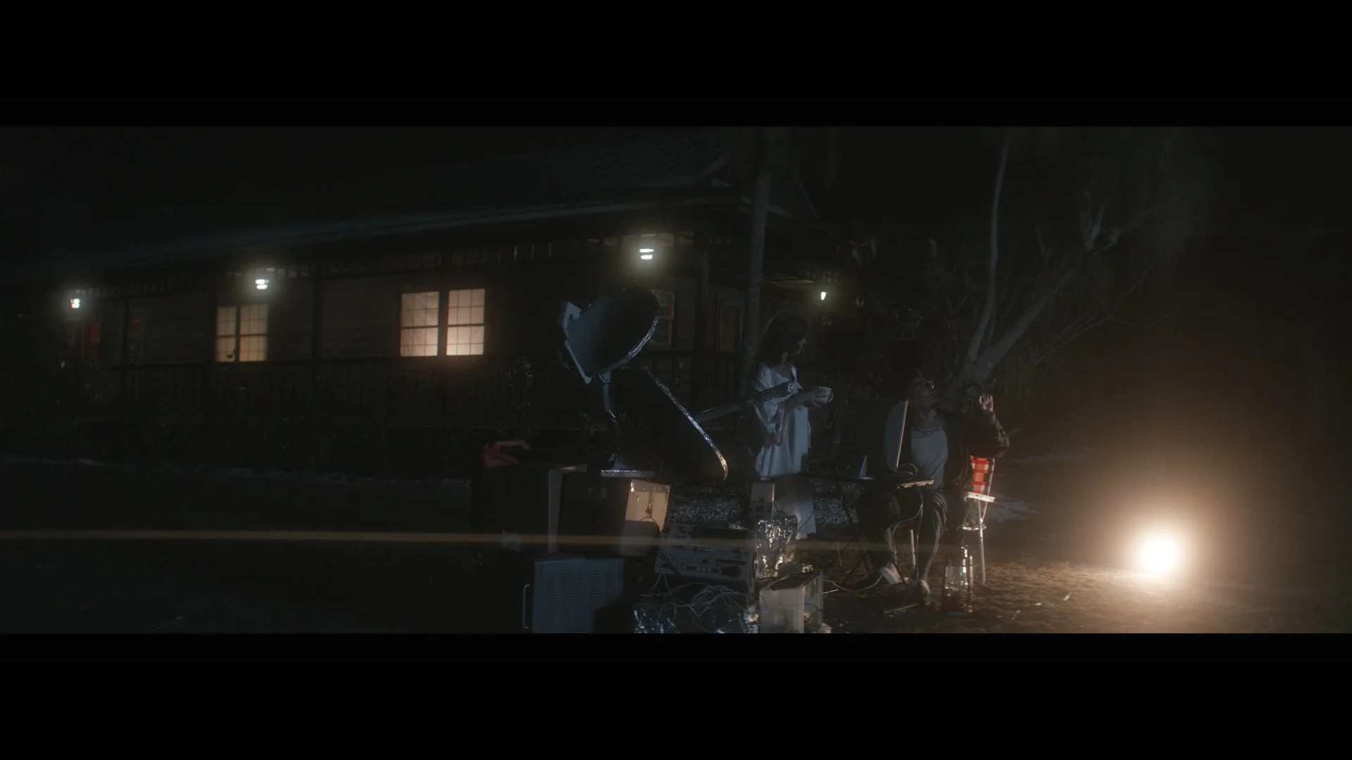 Nighttime outdoor scene with two people sitting on chairs near a bright fire, surrounded by electronic equipment and a large satellite dish. A house with lit windows and a tree are in the background.