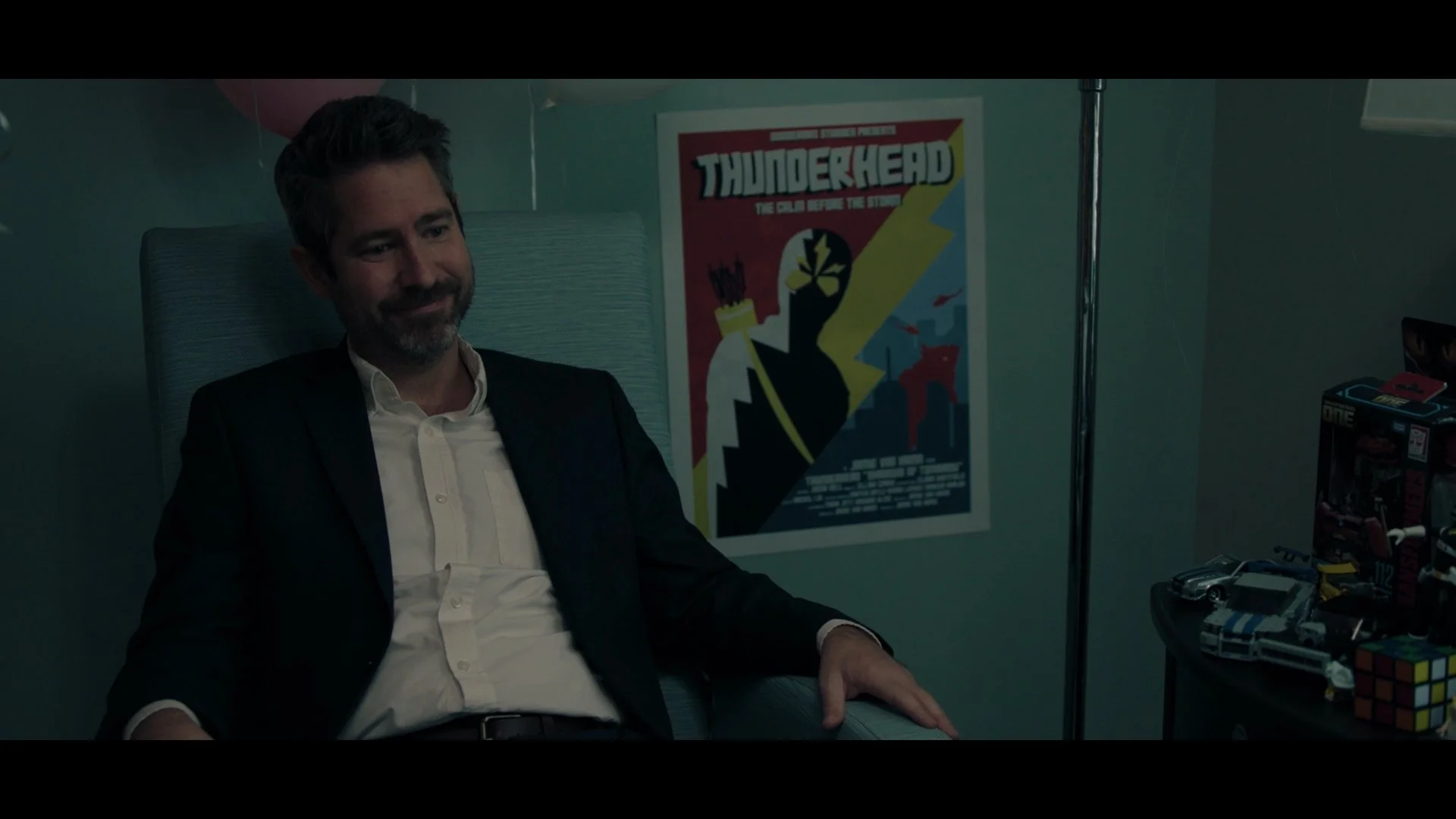 Man in a suit sitting on a chair in a room decorated with a poster and toys, smiling with eyes closed.