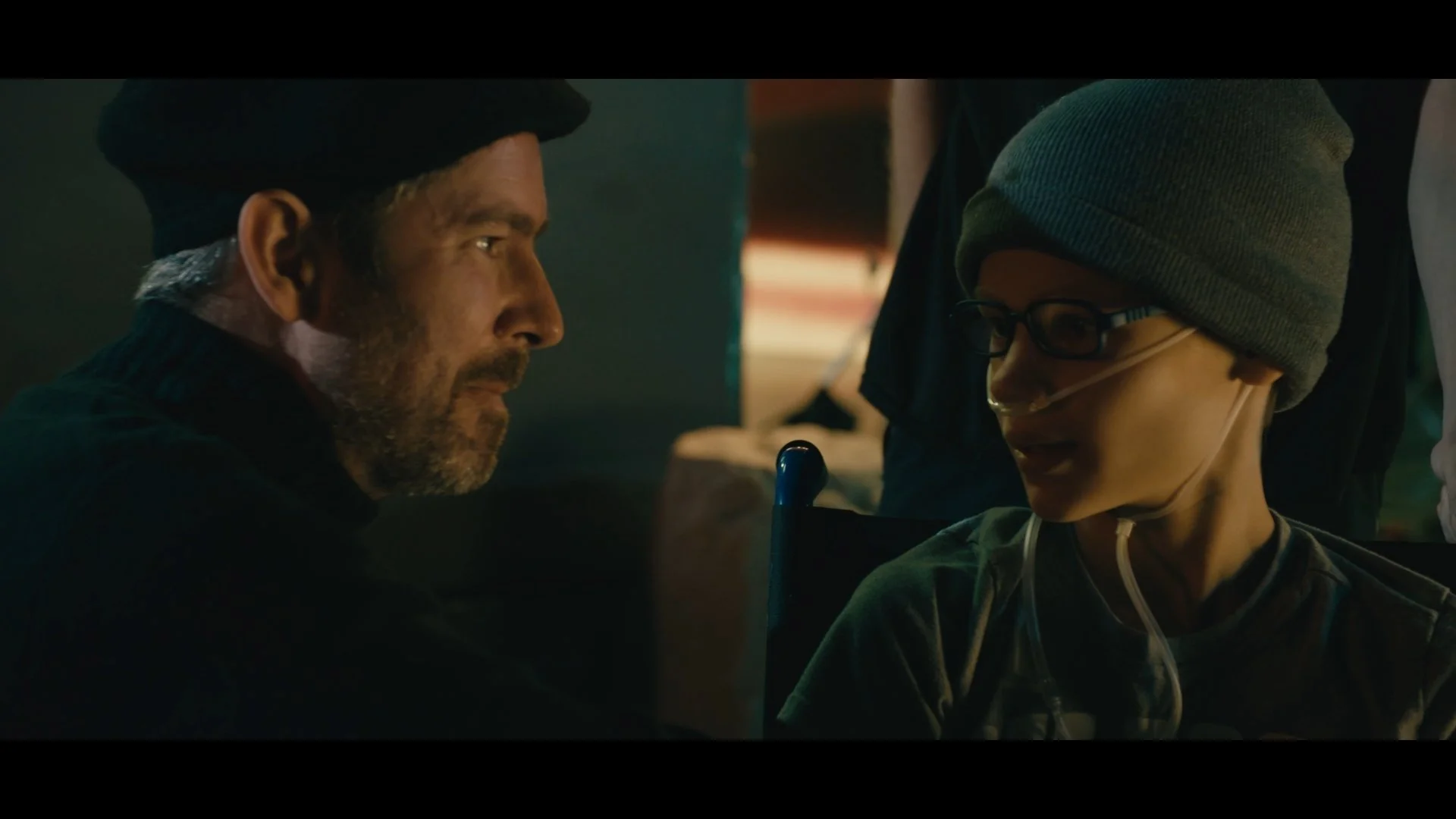 A man with a beard, wearing a black hat, is looking at a young woman with glasses and a grey beanie, who has a nasal cannula, in a dimly lit room.