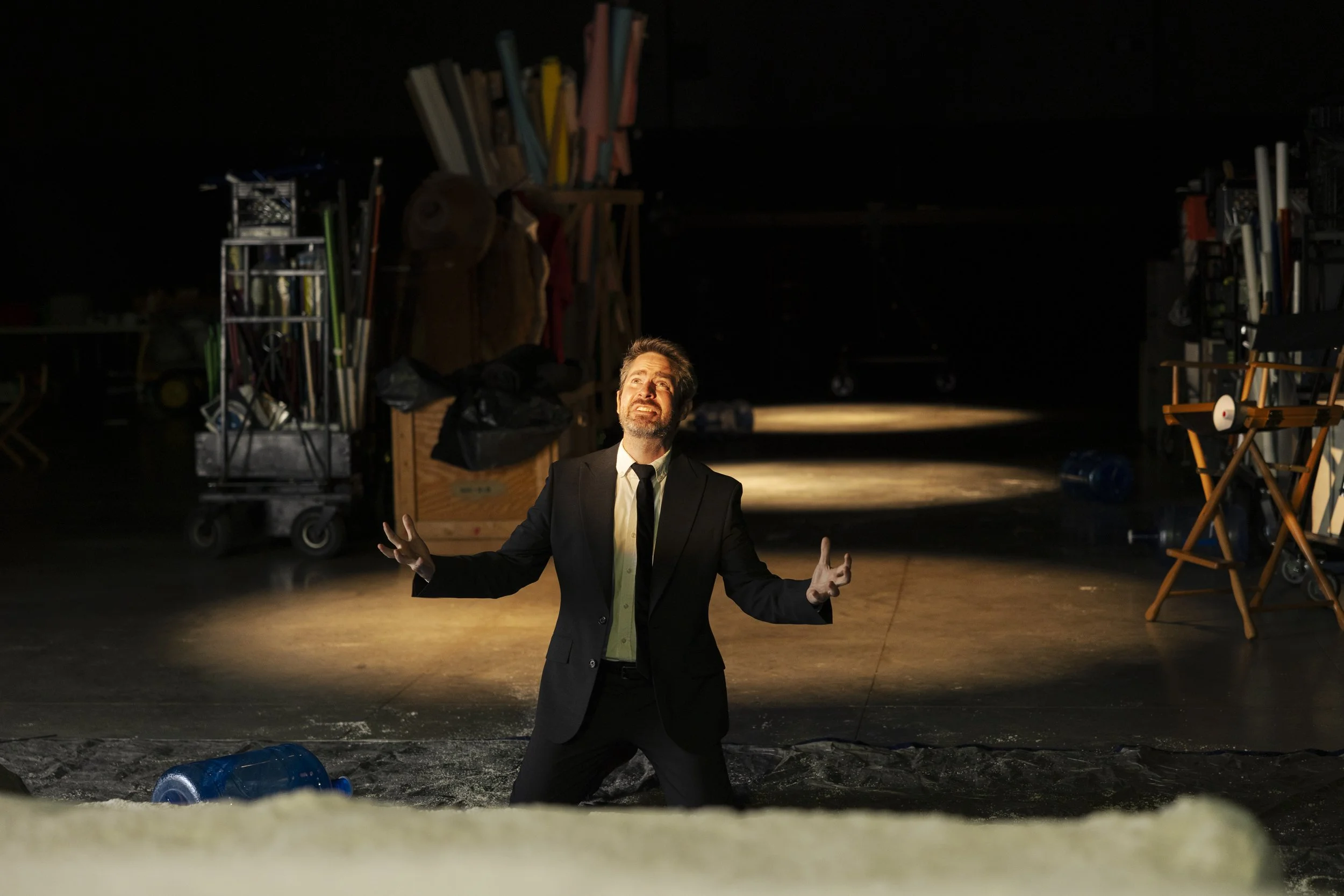 A man in a suit with an expressive face stands with arms open on a stage or theater set, illuminated by a spotlight against a dark background with various equipment and props.