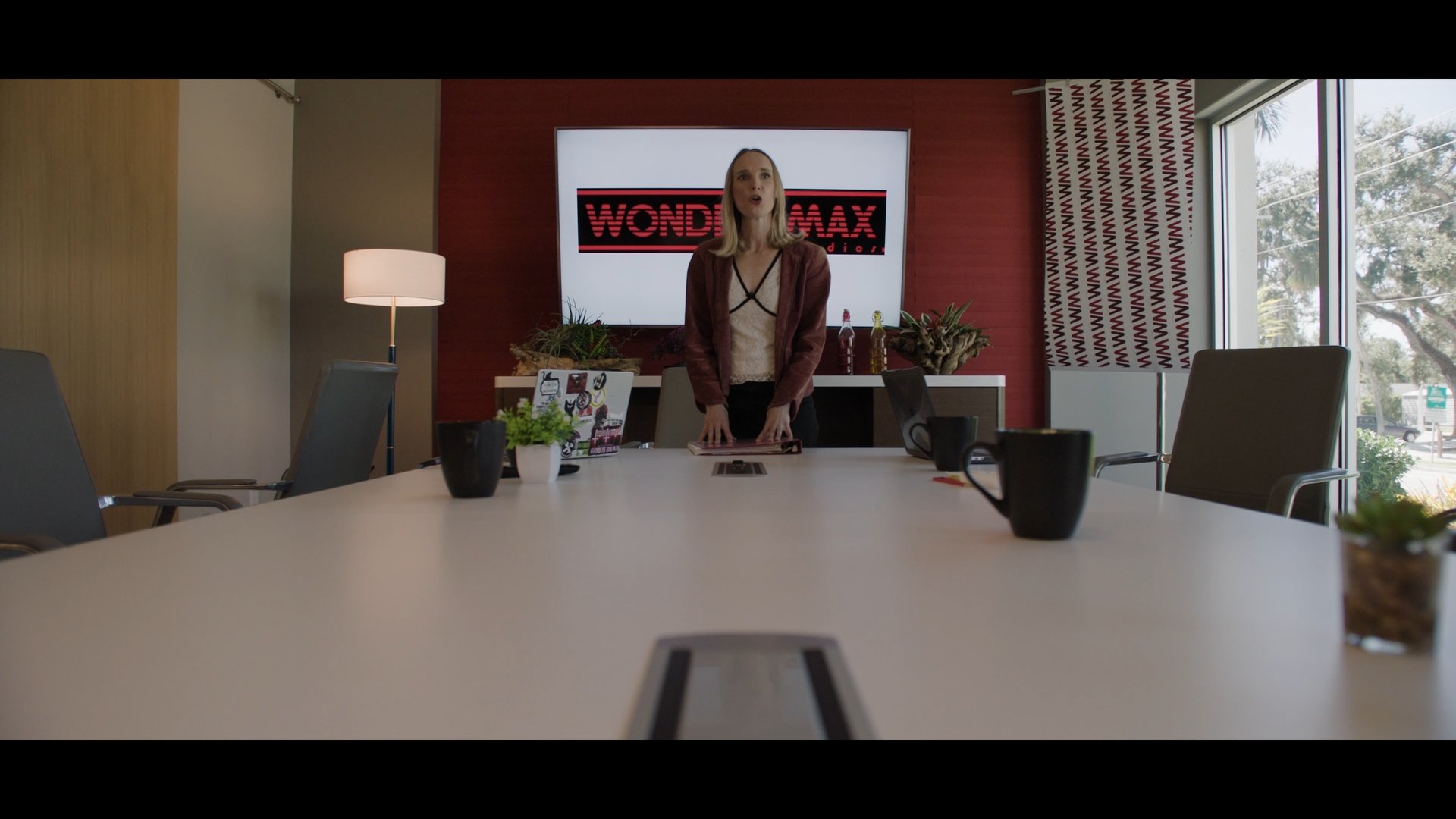 Woman standing at a conference table in a meeting room on a video call, with a WOND MAX logo on a screen behind her.