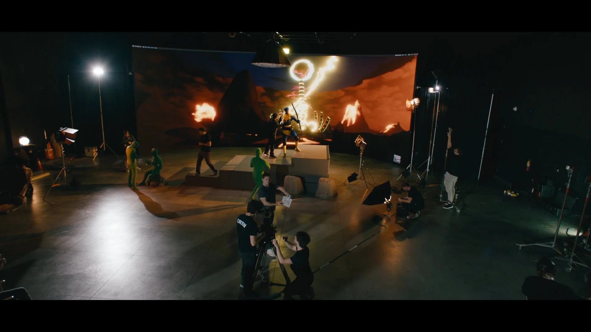Filming studio with a scene of characters in costumes on a set with a volcanic landscape backdrop, crew members operating equipment, and lighting.