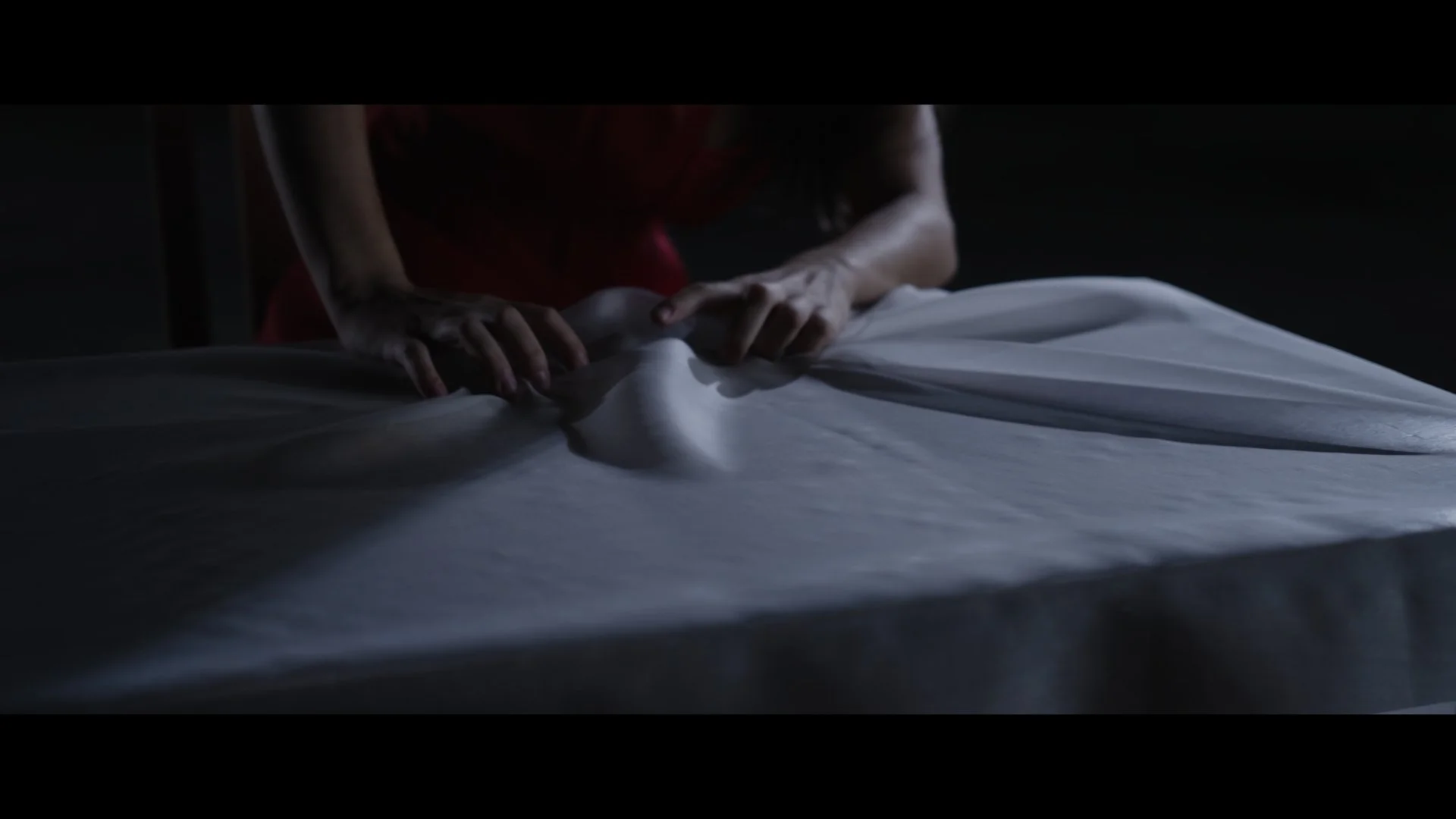 Person leaning over and gripping a white sheet or blanket covering a bed in a dimly lit room.