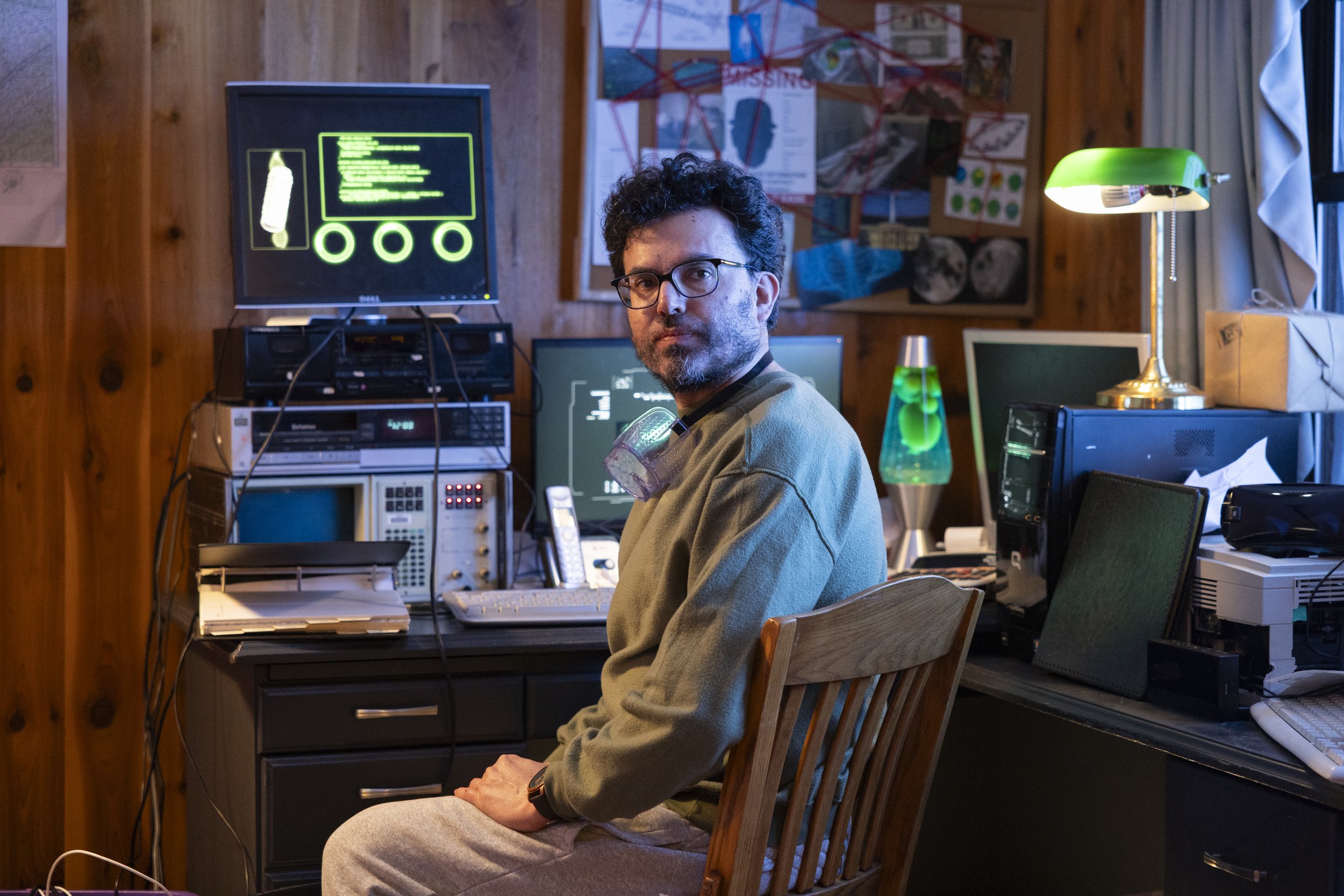 A man with curly hair and glasses sitting at a cluttered desk in a room with wood-paneled walls, surrounded by multiple computer monitors, electronic equipment, and a lava lamp, with various posters and a bulletin board in the background.