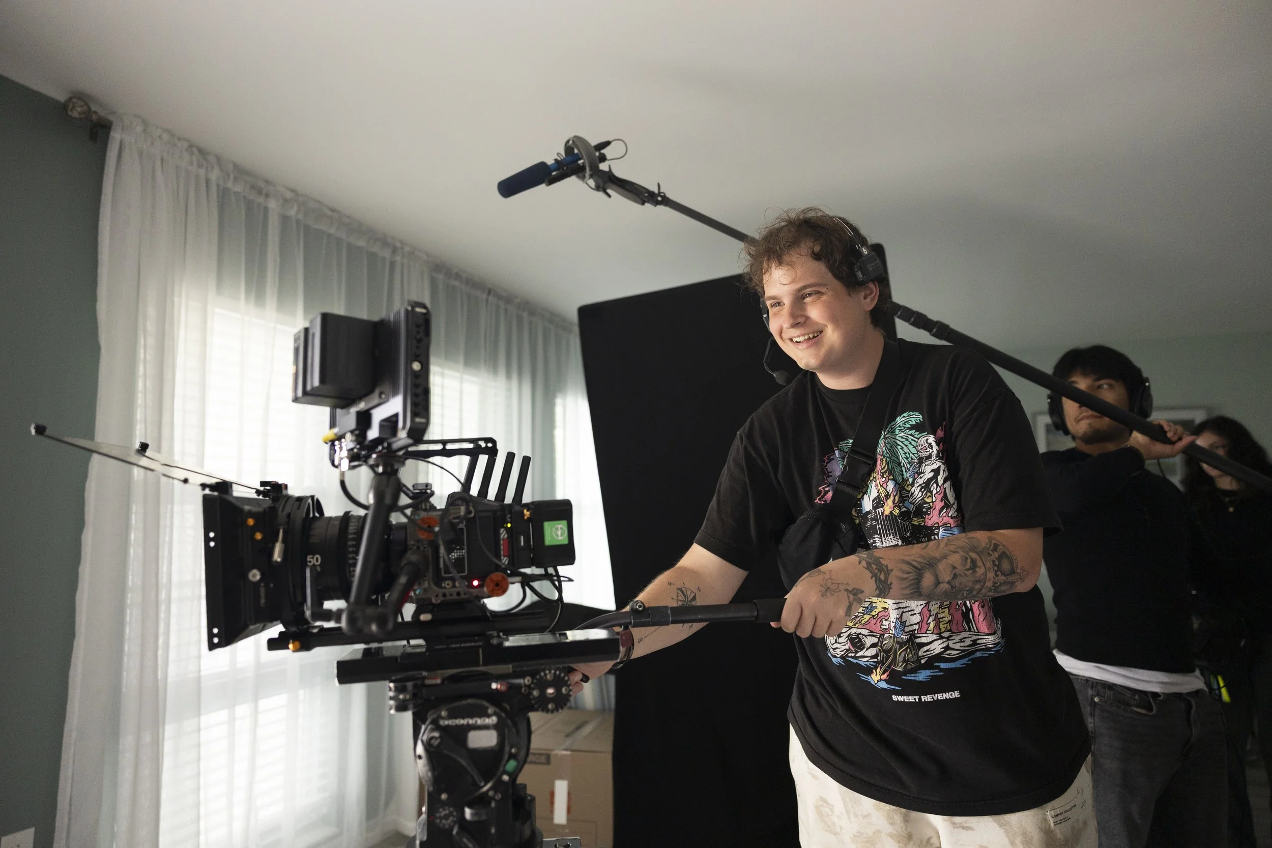 A young man with curly hair smiling while operating a professional video camera setup on a stabilizer in a room with green walls and white curtains.