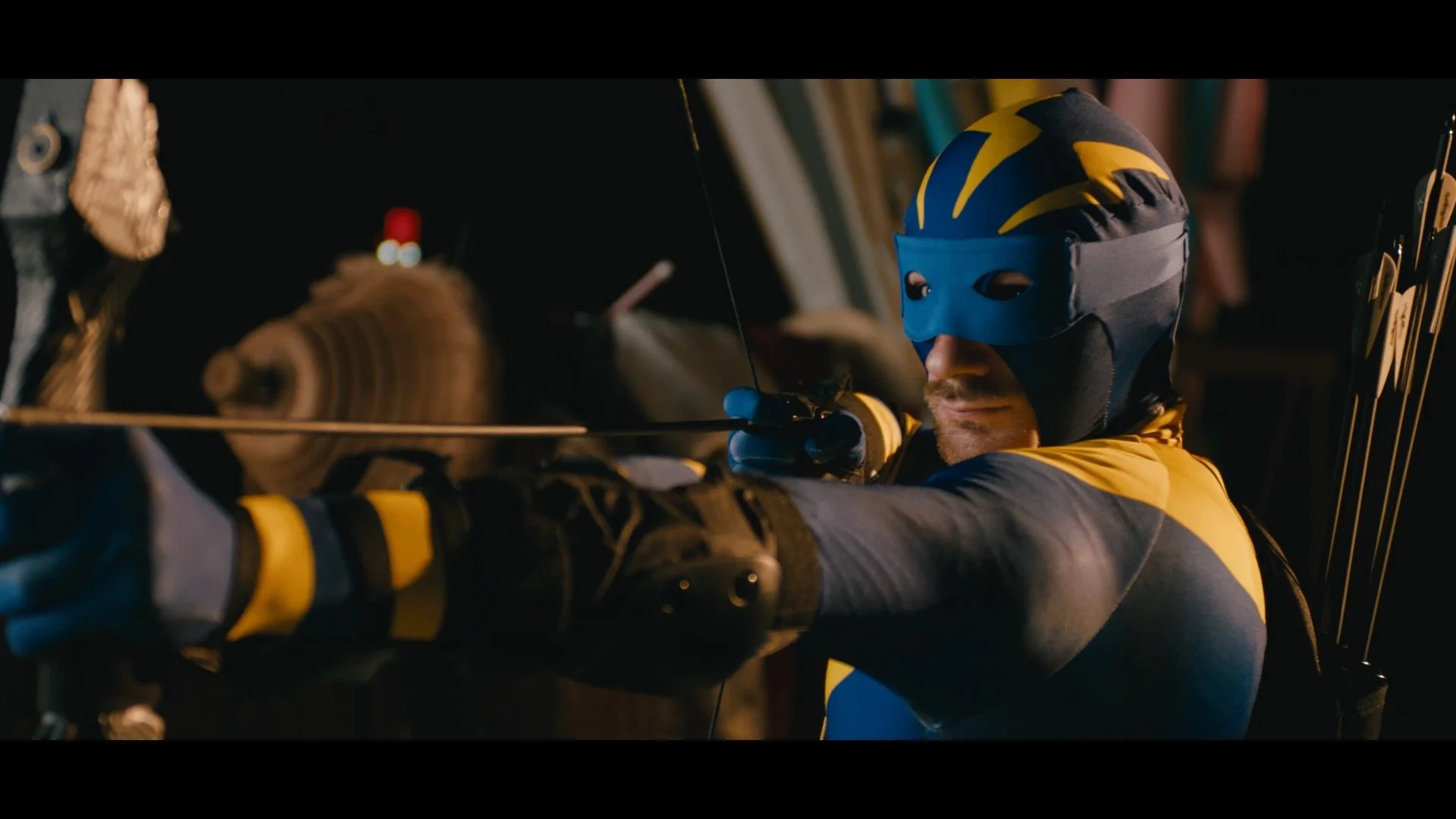 A man wearing a blue and black ski mask and jacket, aiming a bow and arrow in an indoor setting.