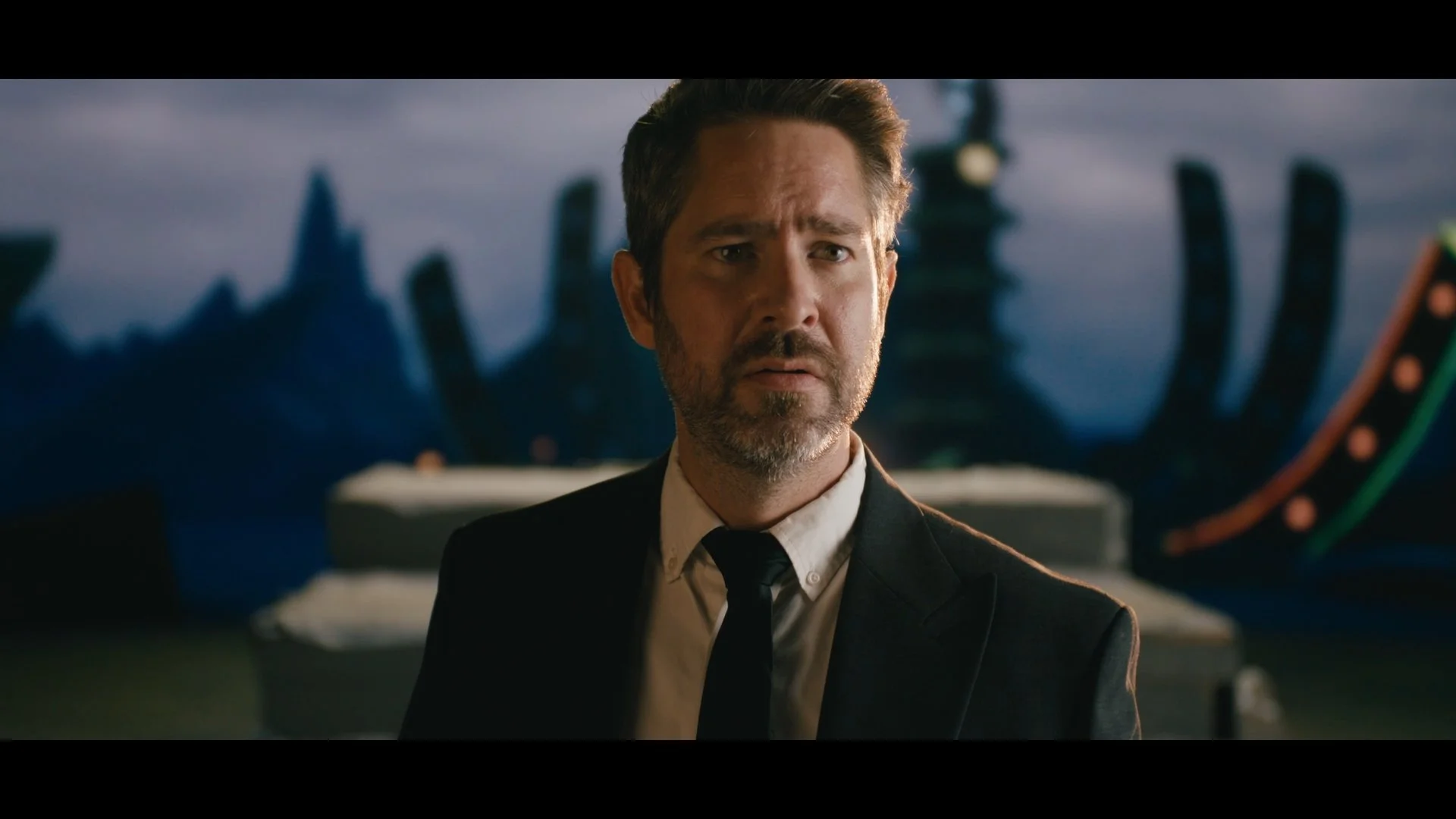 A man in a suit and tie looks concerned or confused in an indoor setting with a dark cityscape and a large hand with a wheel in the background.