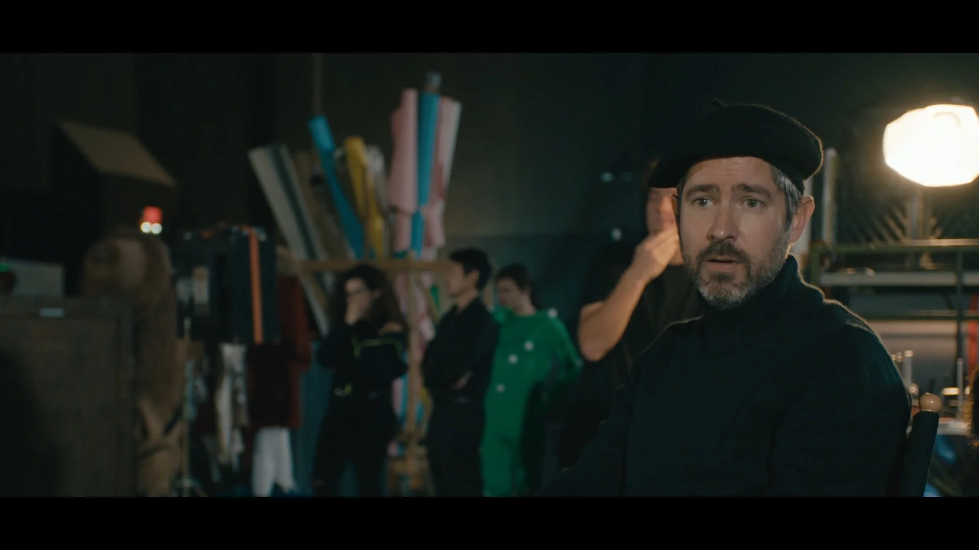 A man with a beard and gray hair wearing a black beret and dark clothing is sitting in an indoor setting with other people in the background. The scene is dimly lit.