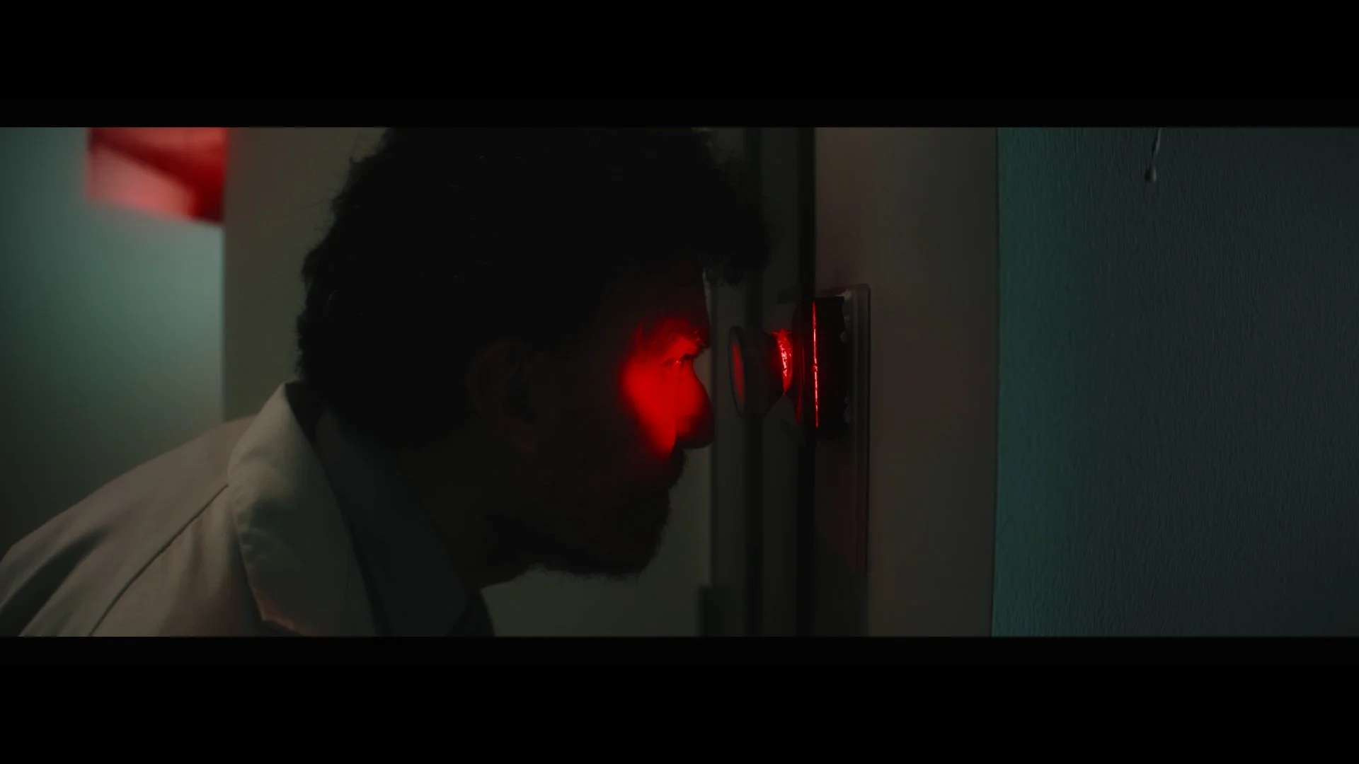 Side profile of a man with dark hair and beard in a dimly lit room, his face illuminated by a red laser-like light coming from a device on the wall.