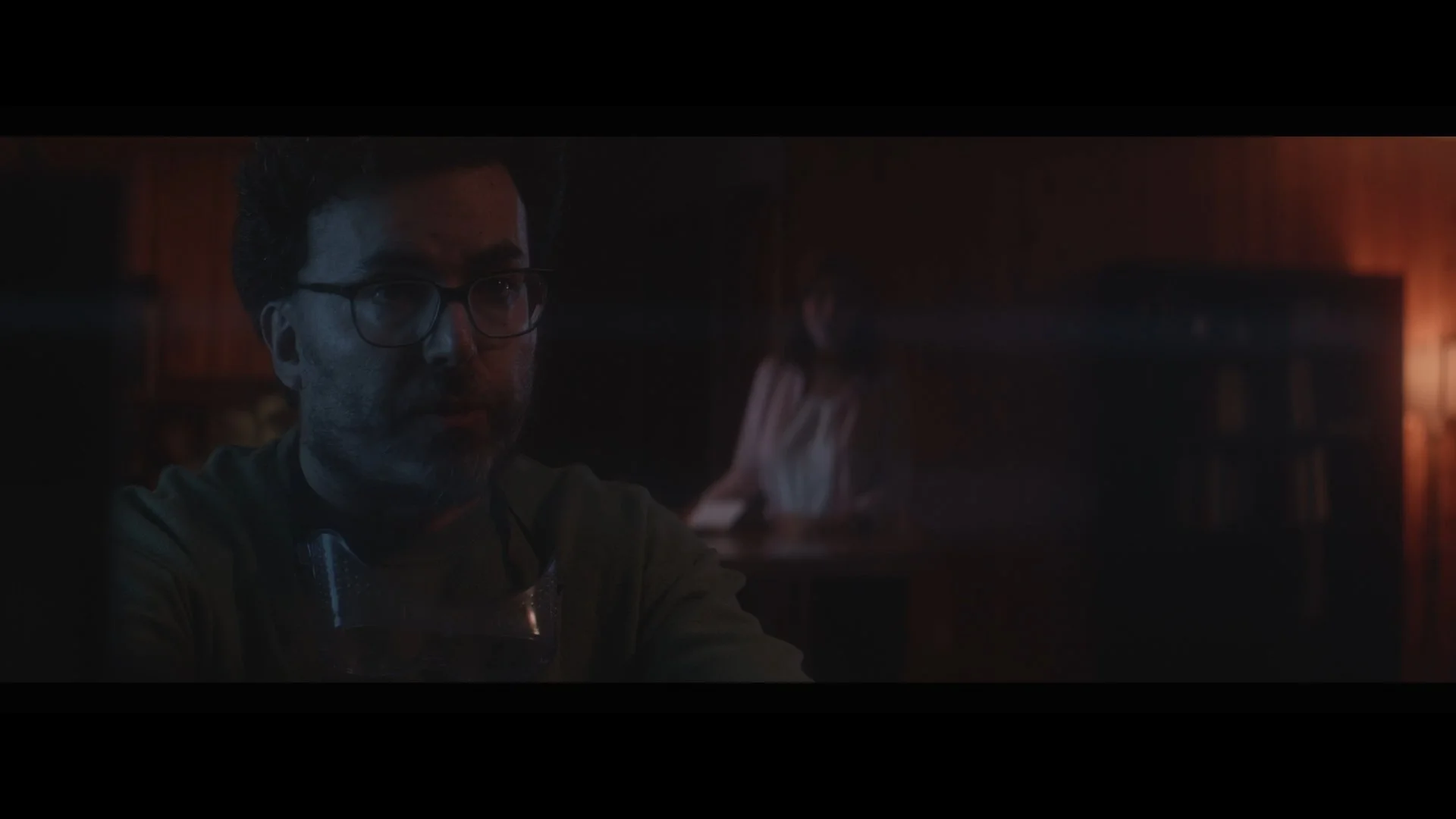 A man with glasses and facial hair looking down, with a woman in the background leaning over a table in a dimly lit room with warm lighting.