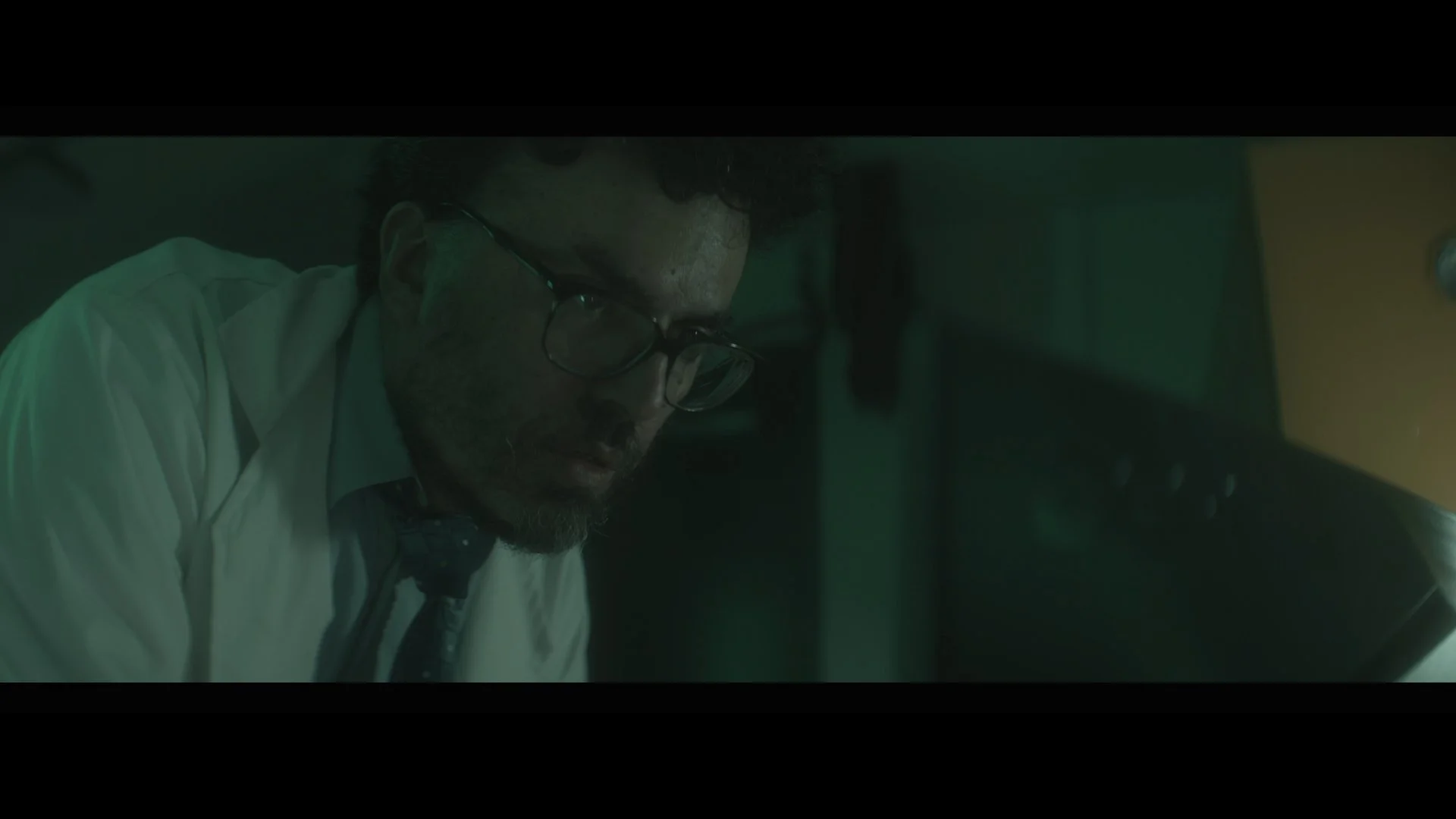 A man with glasses, dark curly hair, and a beard wearing a beige shirt and tie, looking downward in a dimly lit room.