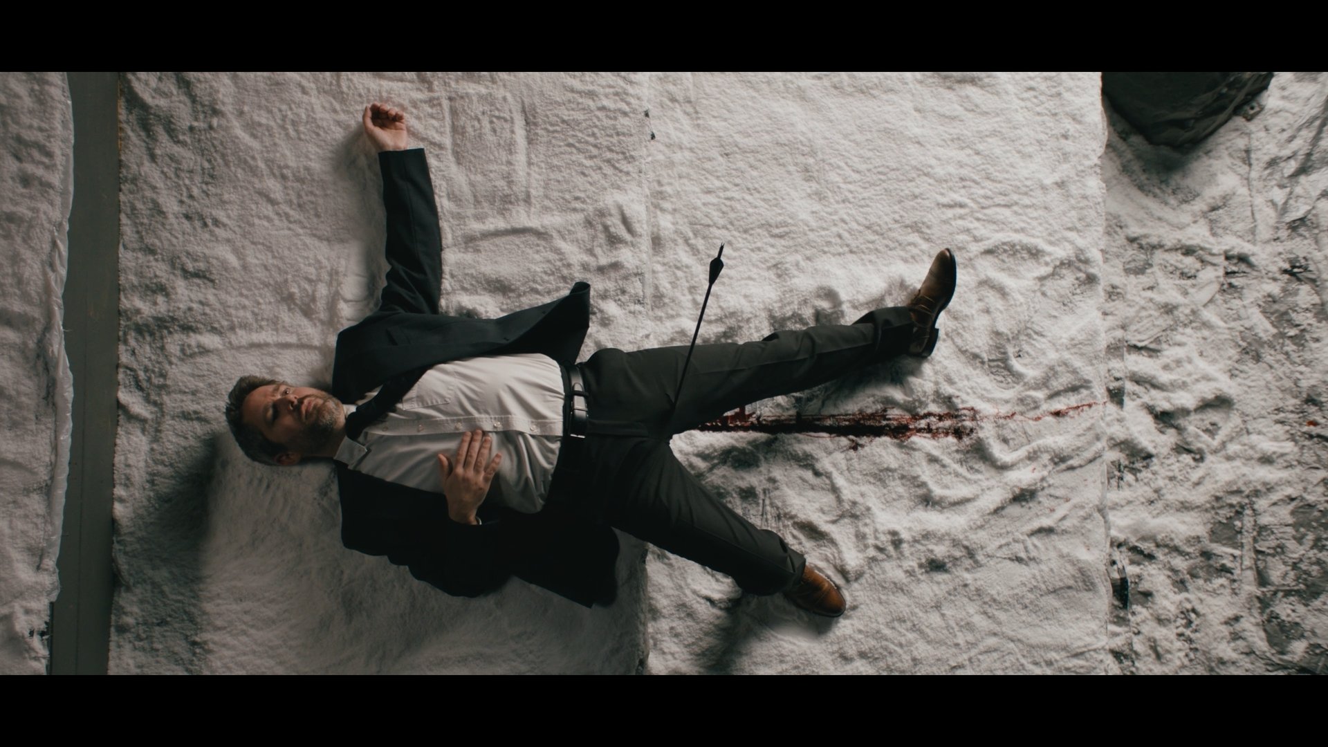 A man in business attire lying on the floor covered in a white powder, with a gunshot wound in his abdomen and blood on the ground.