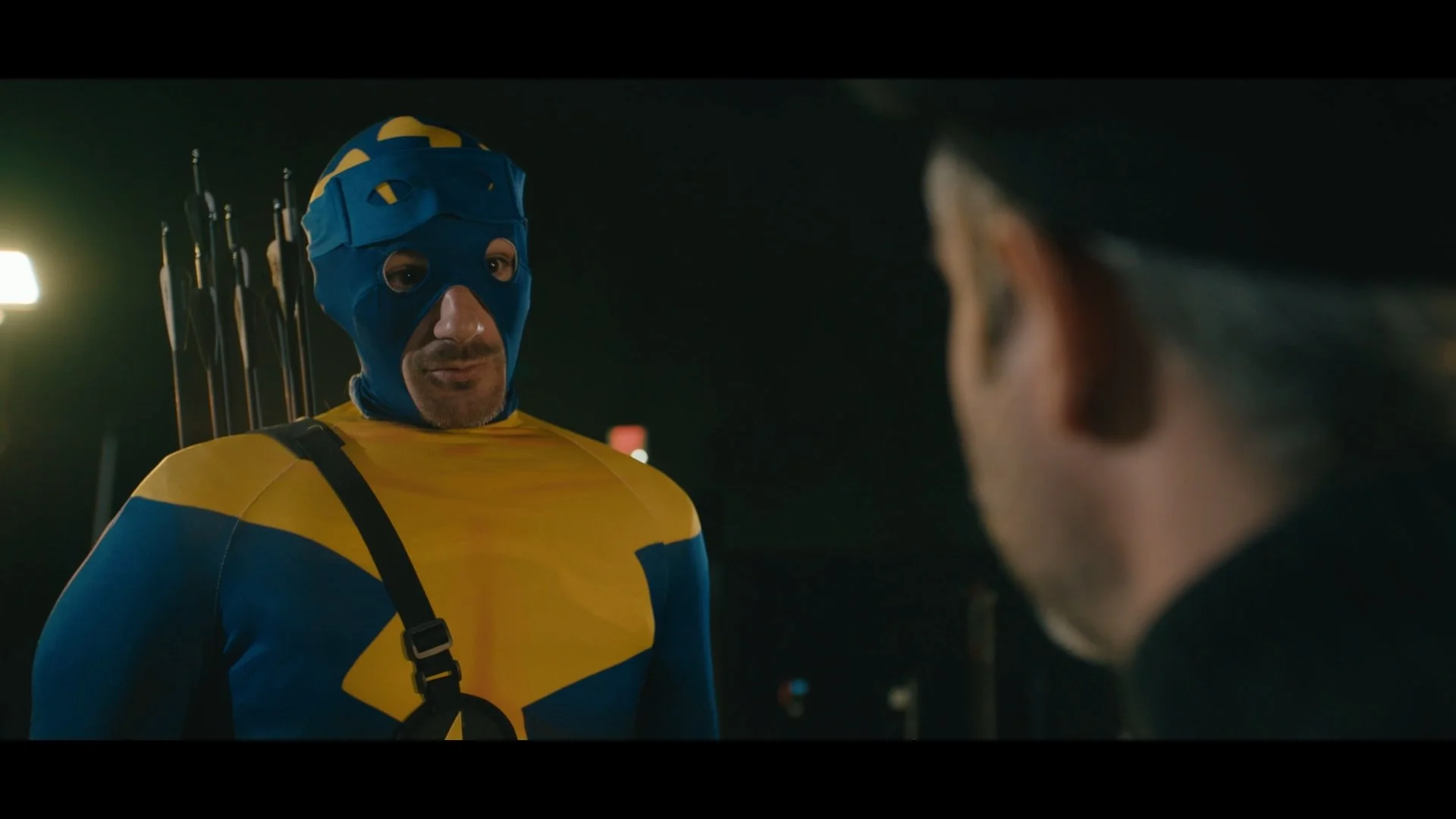 A person dressed as the Swedish superhero Helldiver with a blue and yellow costume, wearing a blue mask and hat, standing at night and talking to another person with grey hair.