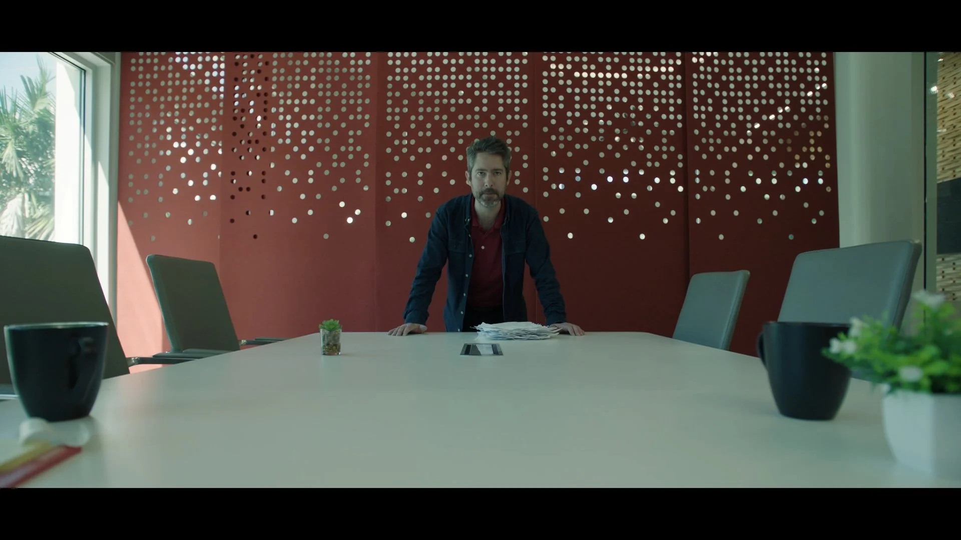 A man standing behind a conference table, leaning forward with hands on the table, surrounded by chairs in a modern room with a decorative perforated red wall and large windows.