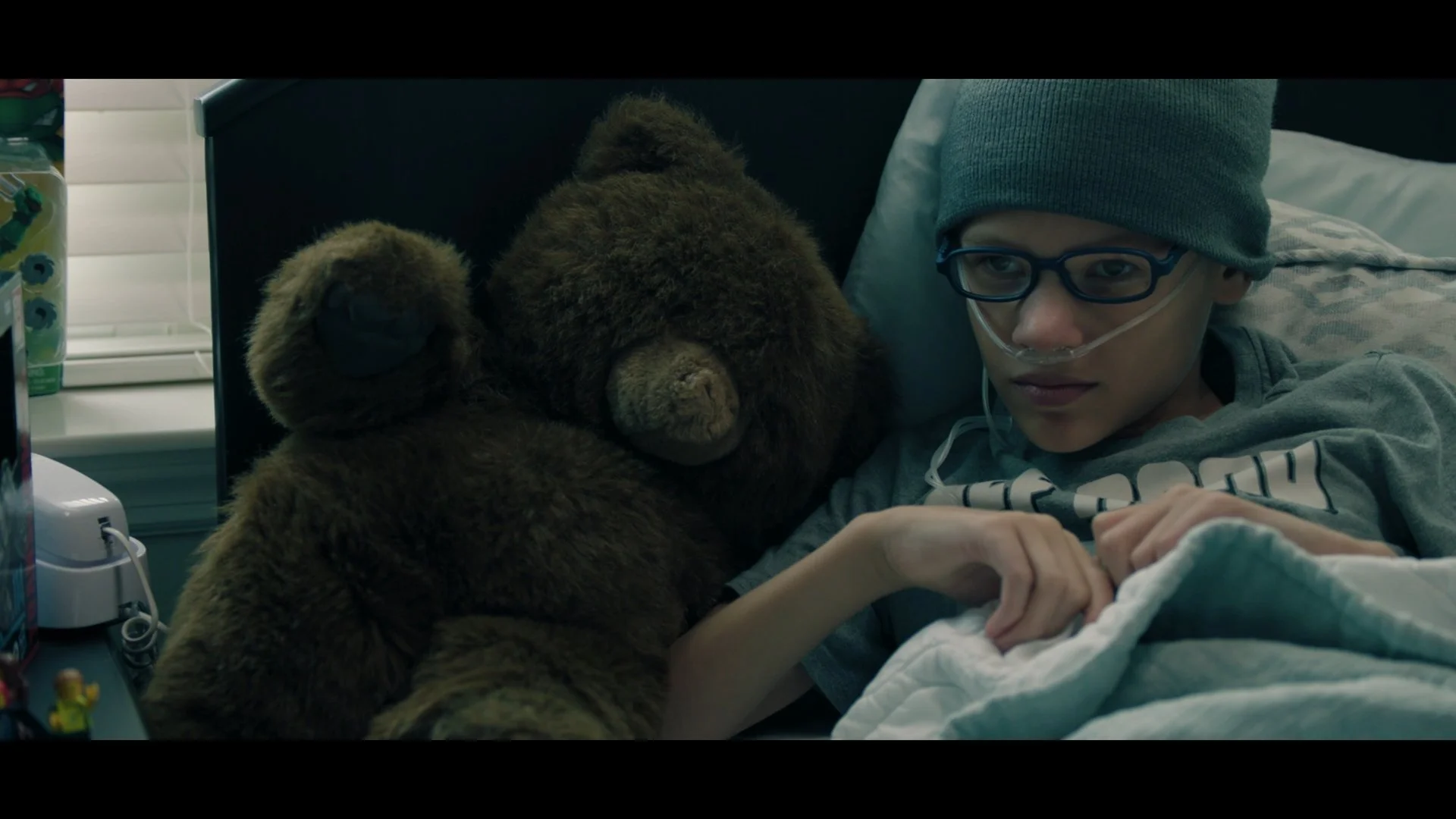 A person wearing glasses, a gray beanie, and a nasal cannula is lying in bed, with a brown teddy bear and a smaller bear beside them.