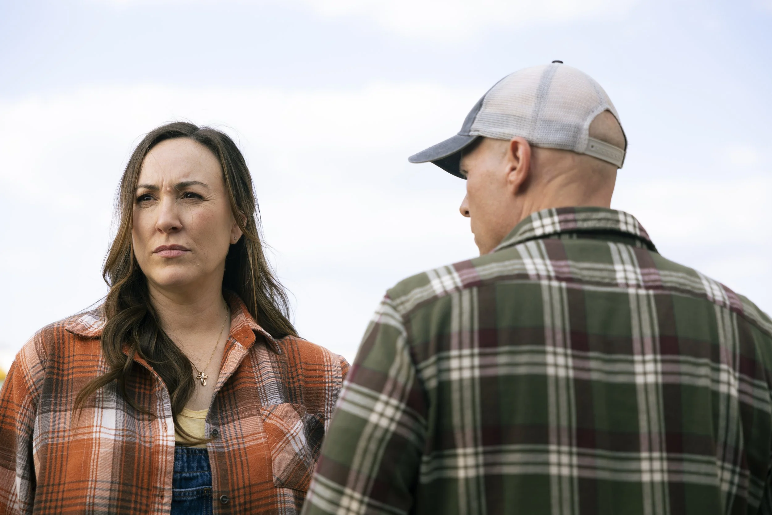 A woman with long brown hair wearing a plaid shirt looks serious while talking to a man with a shaved head, wearing a baseball cap and a checkered shirt, against a cloudy sky background.