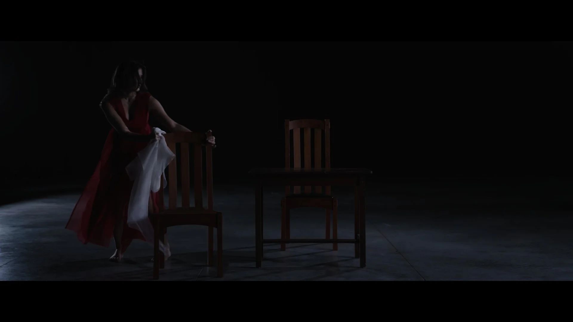A woman in a red dress holding a white cloth, standing next to two wooden chairs and a table in a dark, empty room.