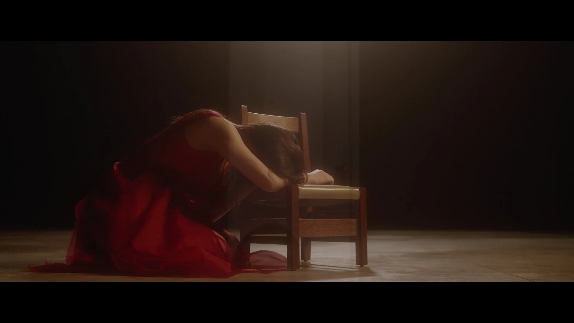 Woman in a red dress kneeling on the floor, resting her head on a small wooden chair in a dimly lit room.