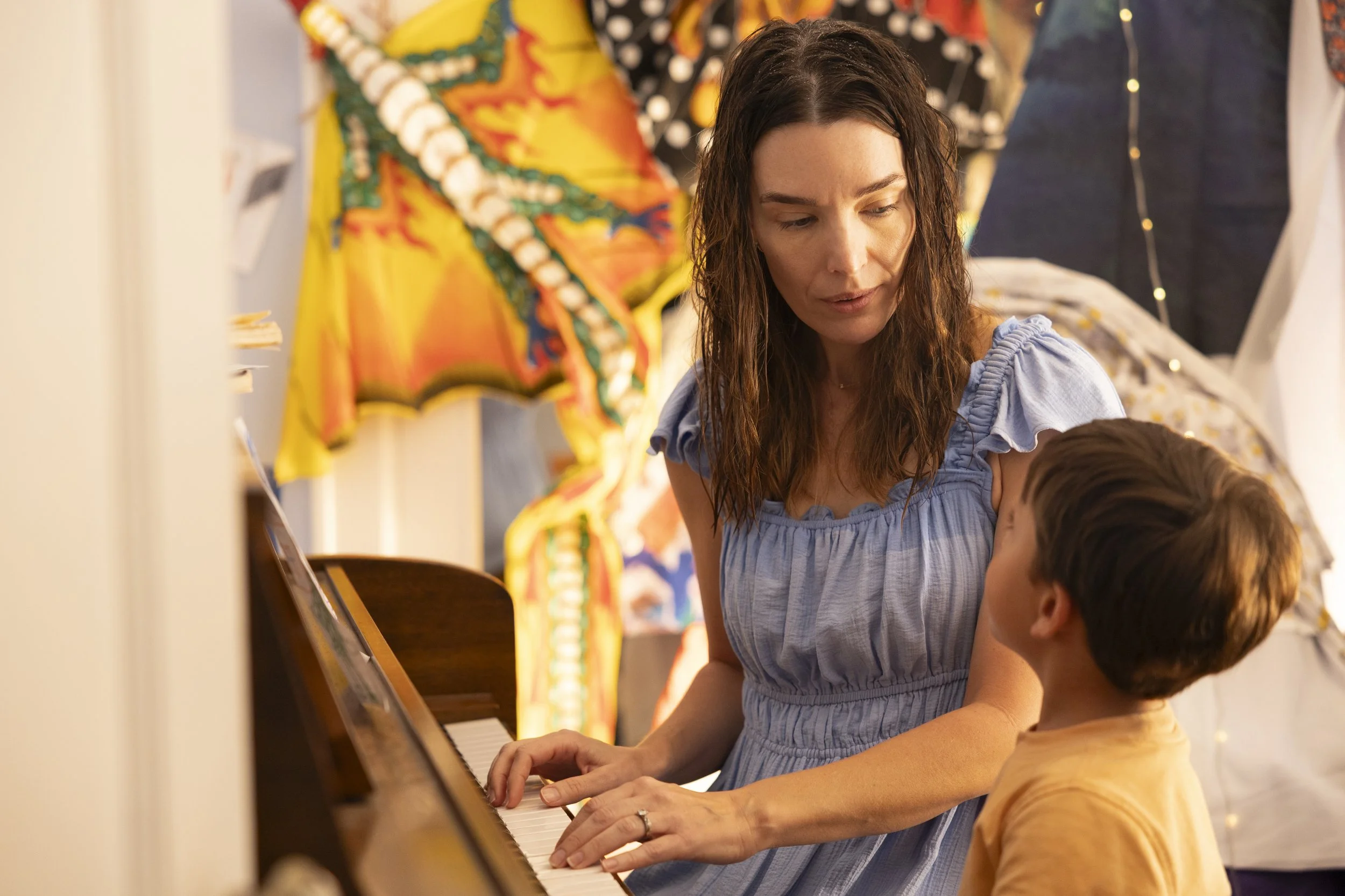 A woman teaching a young boy to play piano in a room decorated with colorful artwork and string lights.