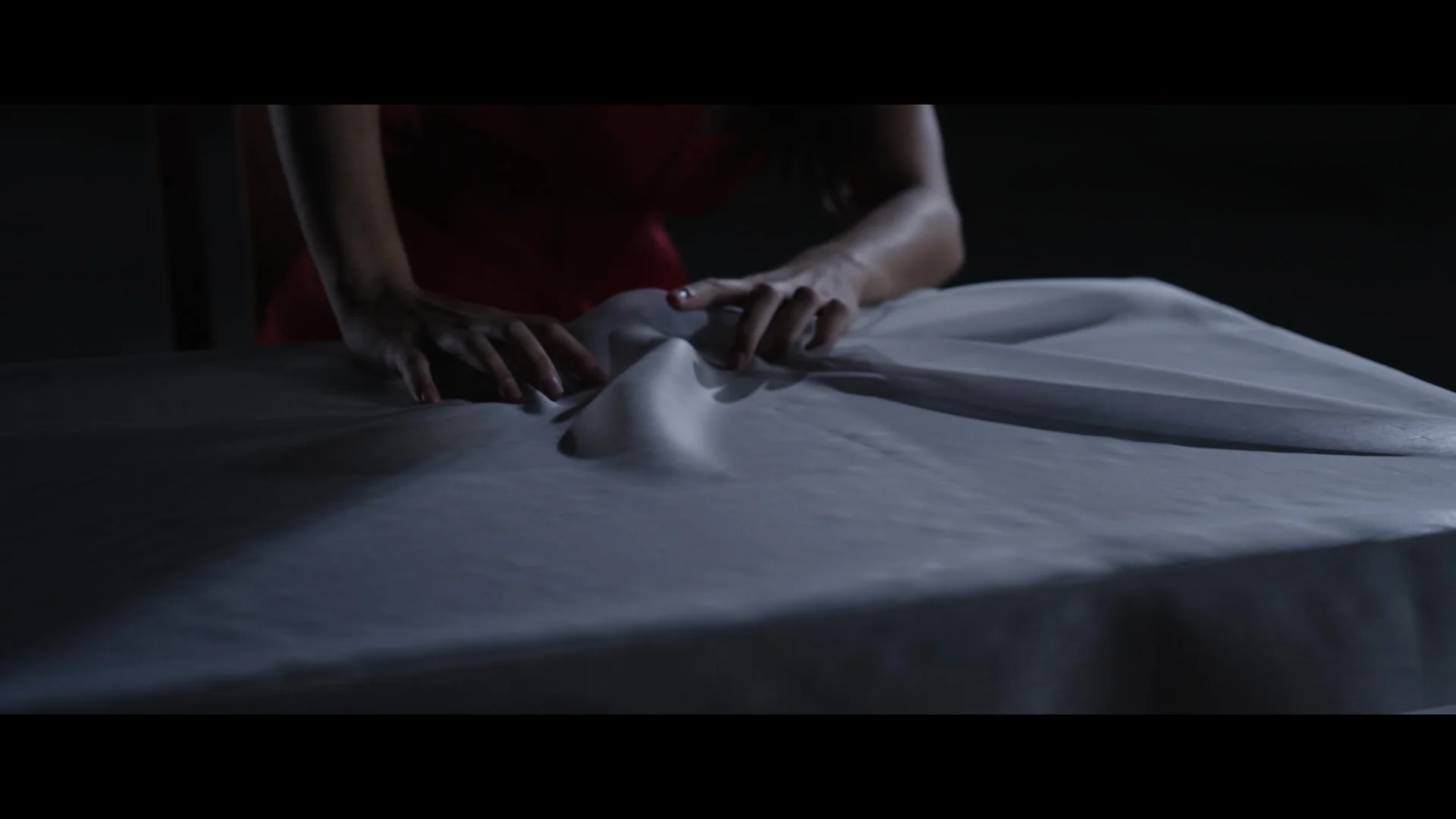 Person in a red dress leaning over a bed with white sheets in a dimly lit room.