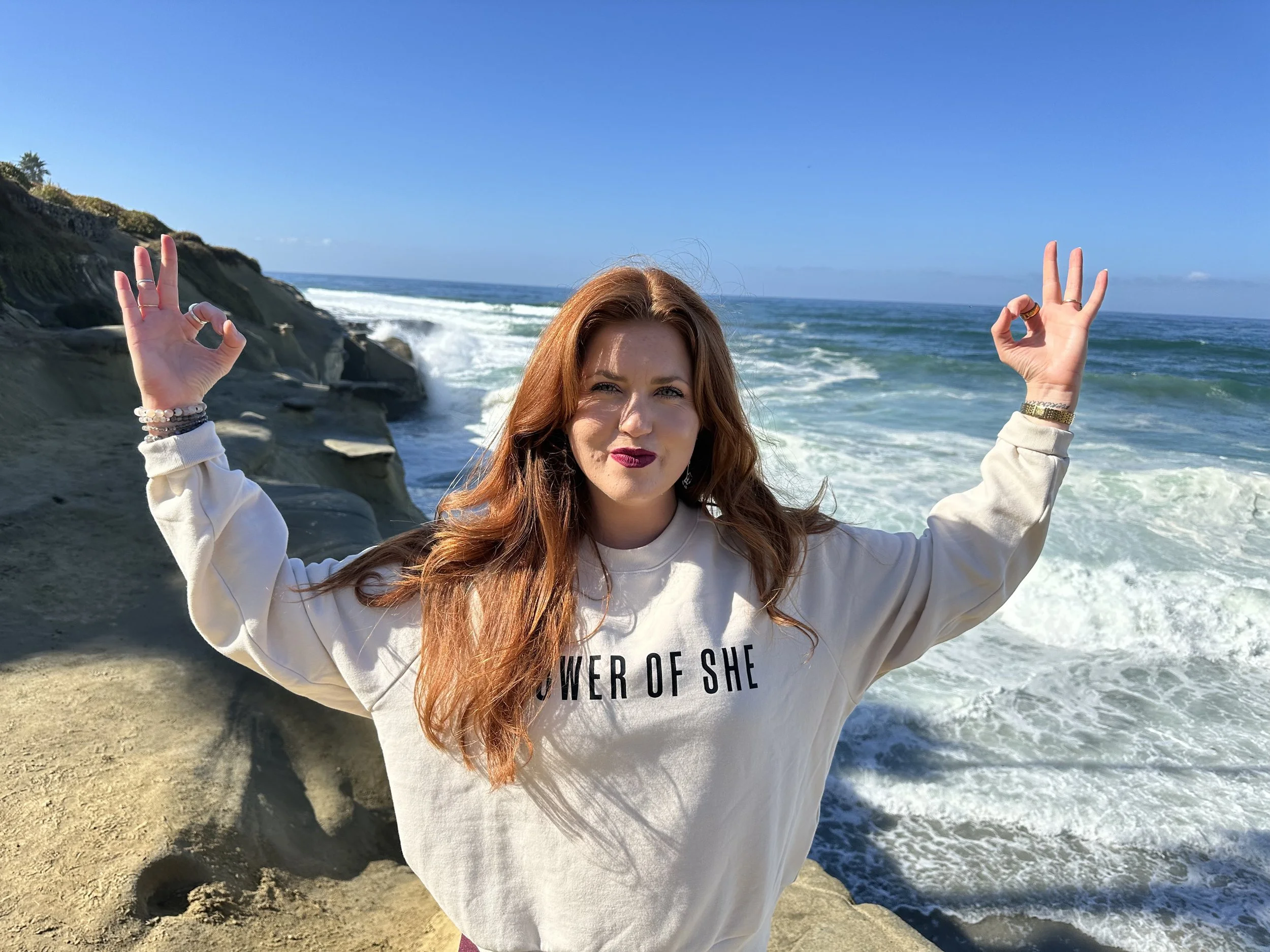 A woman with red hair and dark lipstick standing on a beach, making an OK hand gesture with both hands, with ocean waves and cliffs in the background.