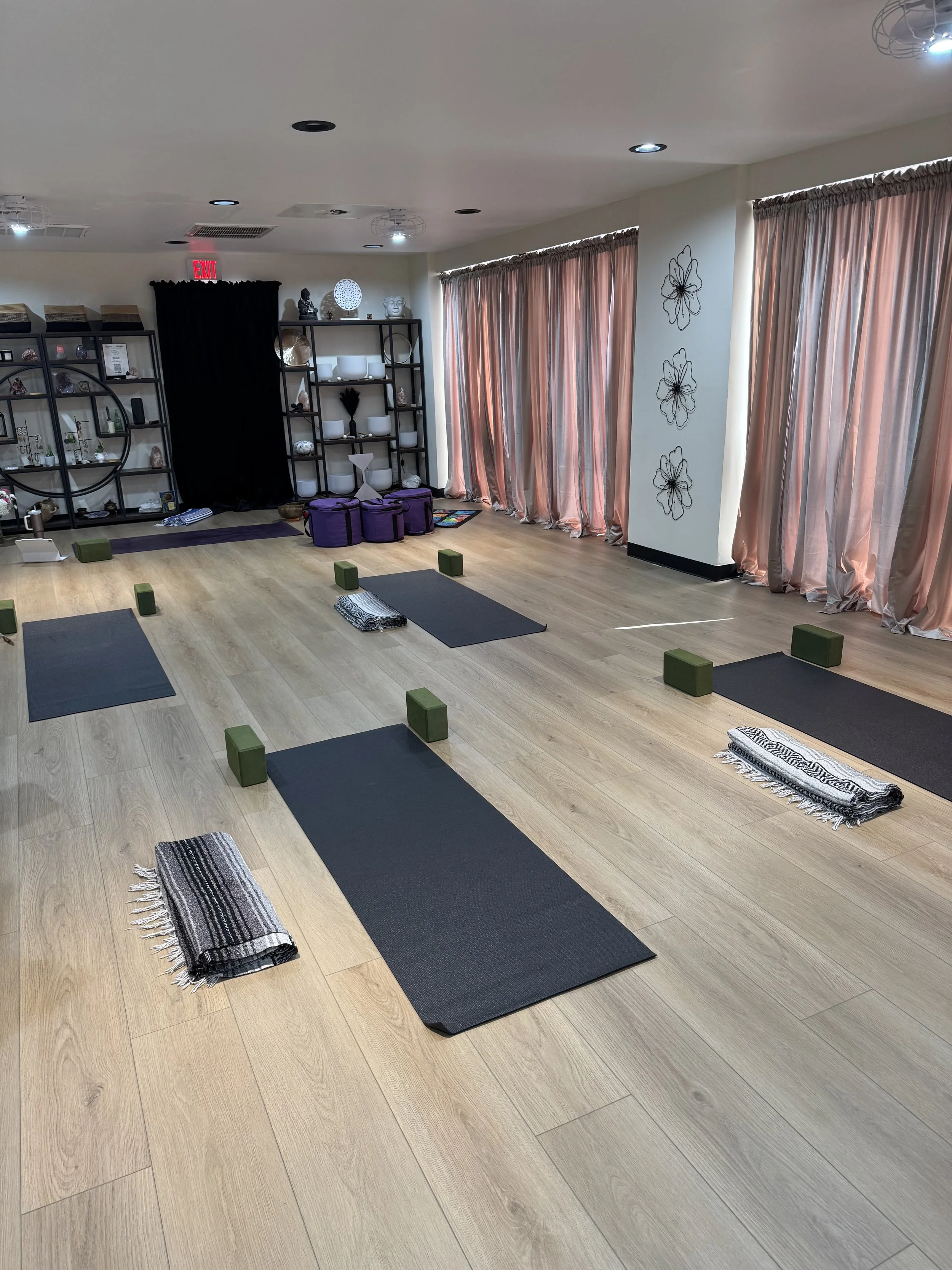A yoga or meditation studio with black exercise mats, green yoga blocks, rolled black and white striped towels, a black curtain, and open shelving with decorative items, sculptures, and plants. Sunlight filters through peach-colored curtains on windows along one wall.