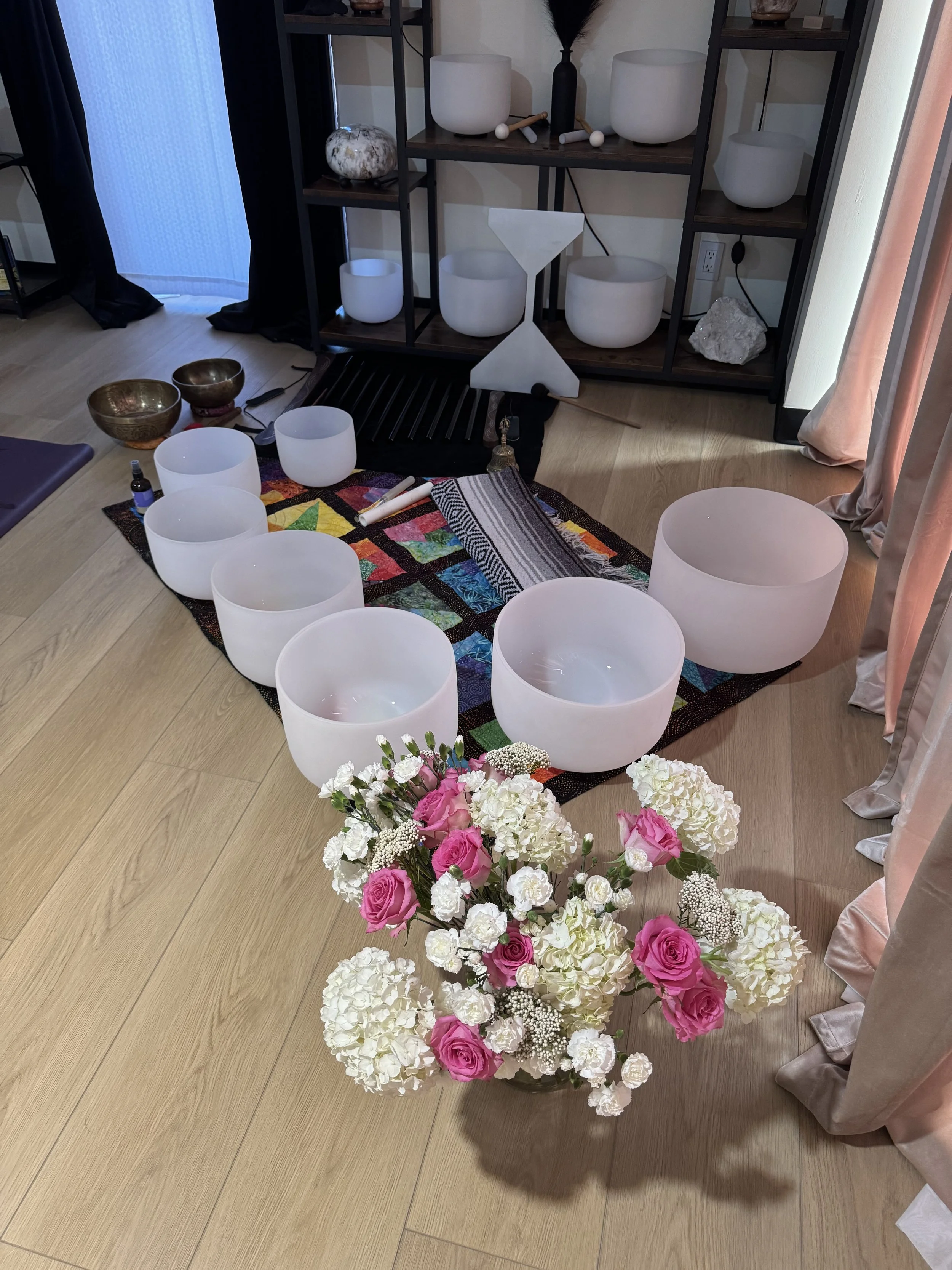 A white and pink flower arrangement on a wooden floor with bowls, maracas, and a colorful quilt nearby, and a black shelf with white bowls, a stone, and decorative objects in the background.