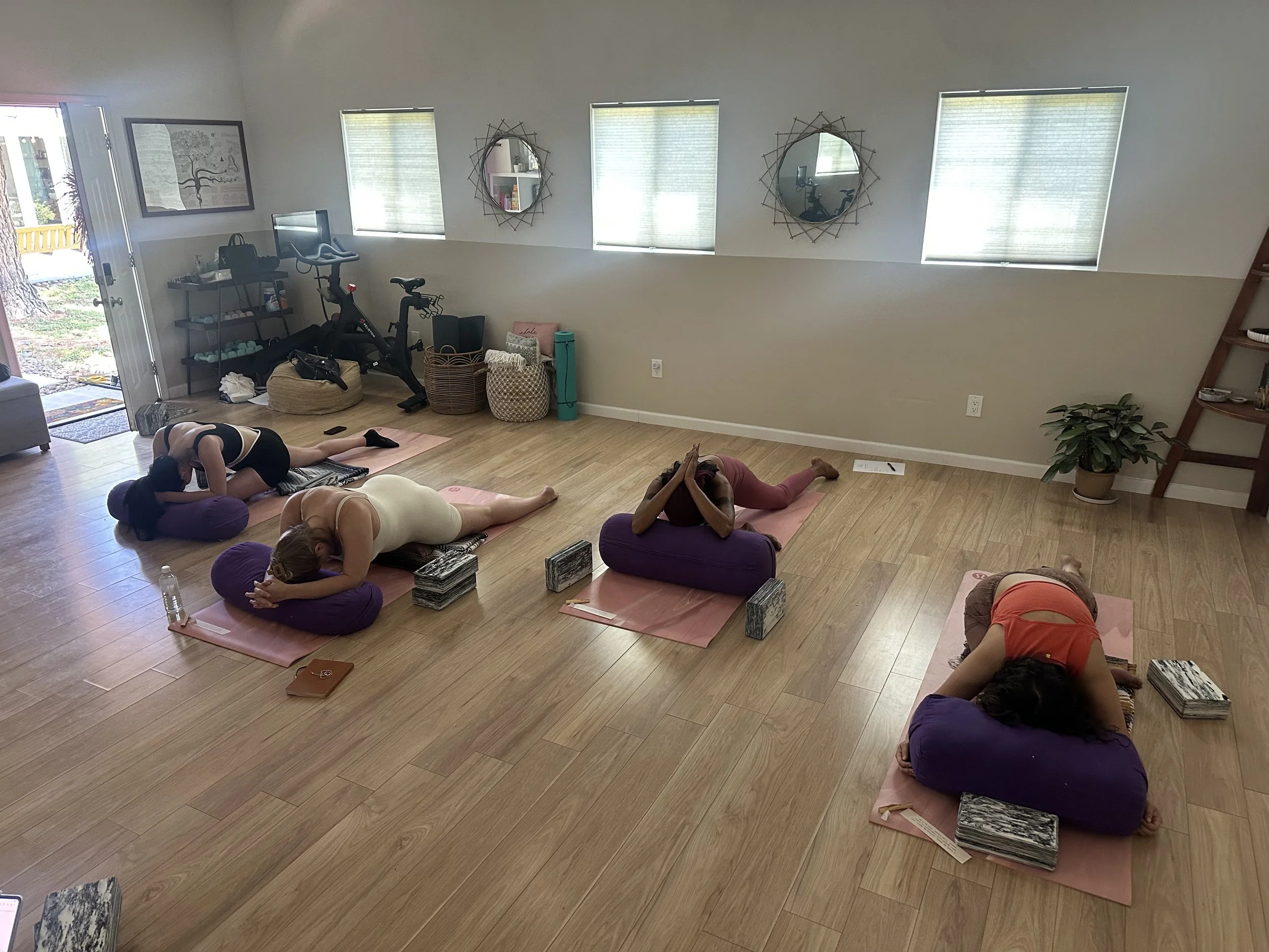 A group of people practicing yoga in a room with wooden floors and three windows, with some using yoga mats and cushions, while mirrors and plants are also visible.