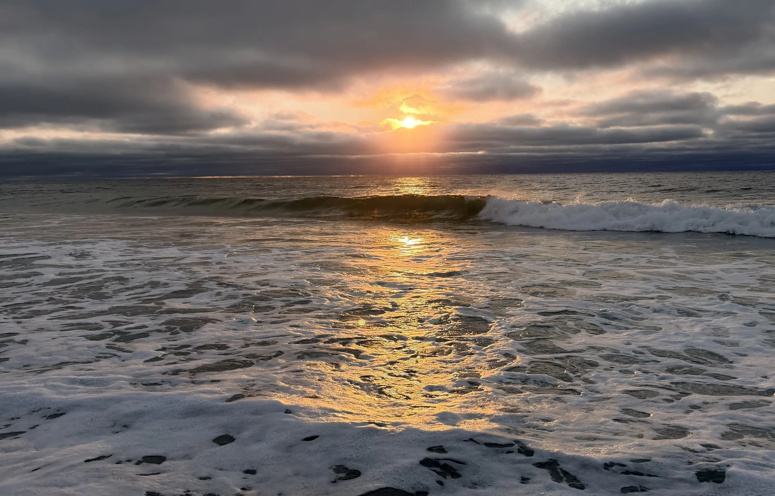 Sun setting over the ocean, clouds in the sky, waves rolling onto the sandy beach, golden reflection on the water.
