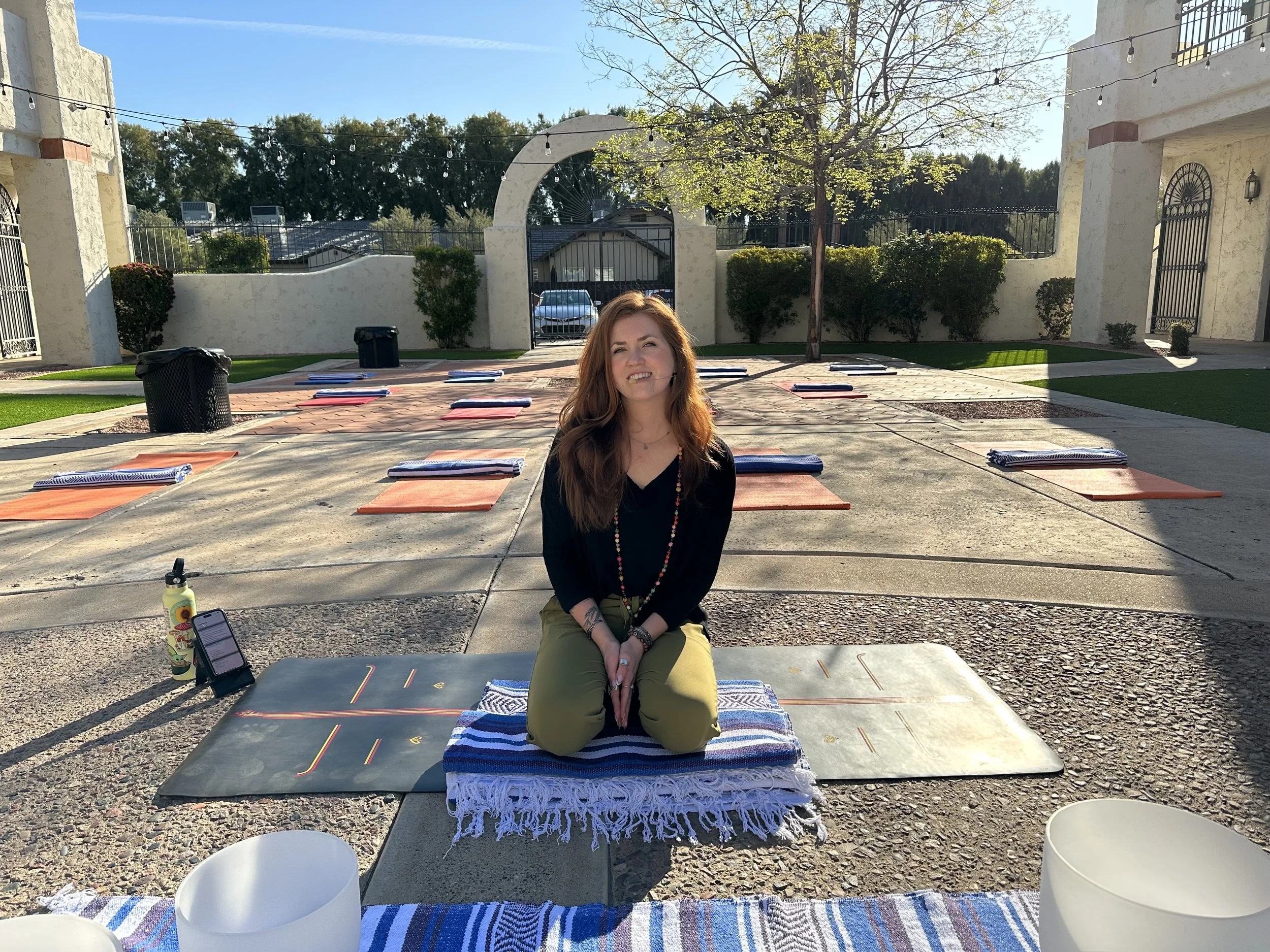 A woman with long red hair practicing yoga outdoors on a sunny day, sitting cross-legged on a yoga mat with a colorful towel on top, amid mats and props set on a patio surrounded by greenery and white stucco buildings.