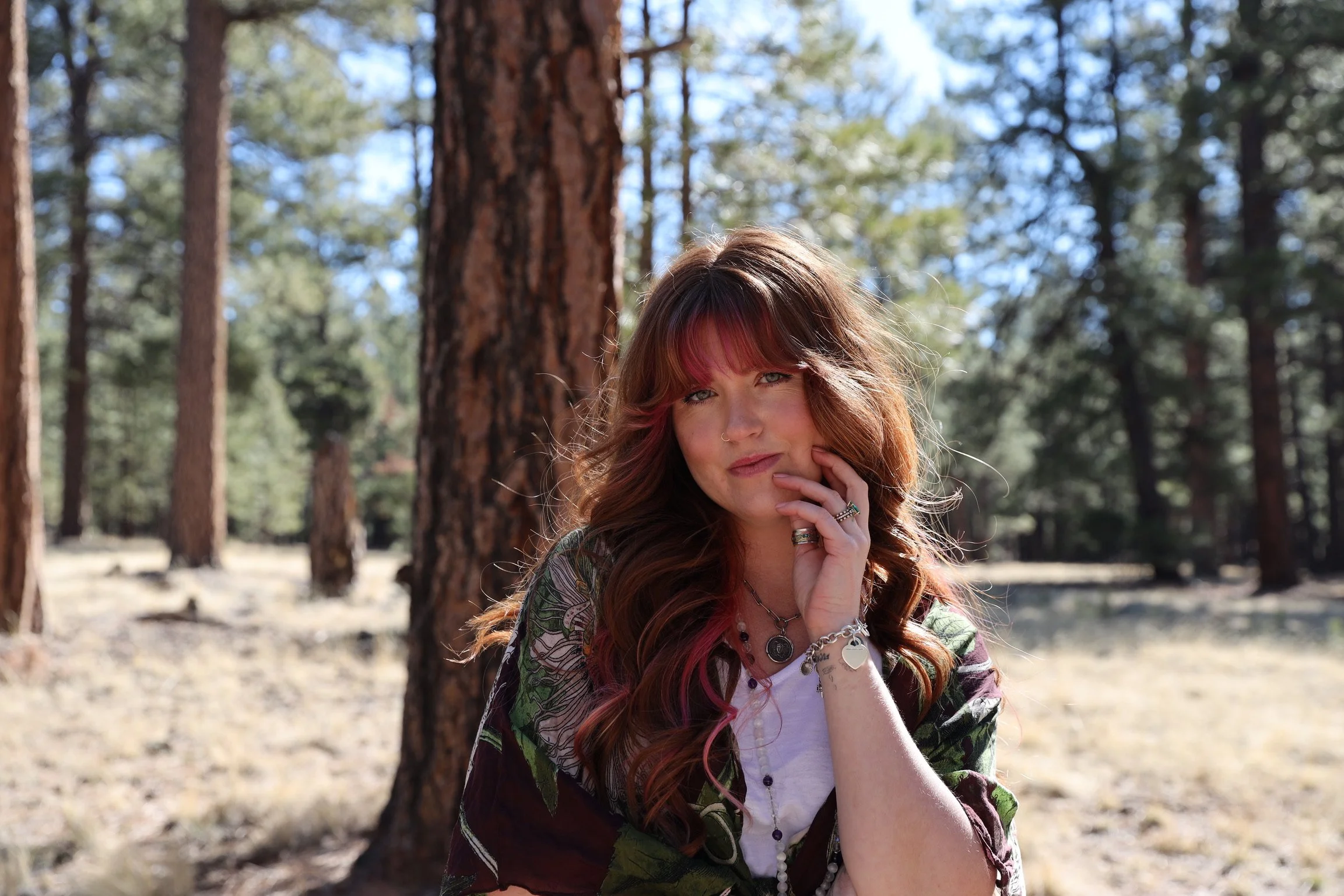 A woman with long red hair and colorful streaks, wearing rings, necklaces, and a patterned shawl, posing outdoors near trees in a forested area.