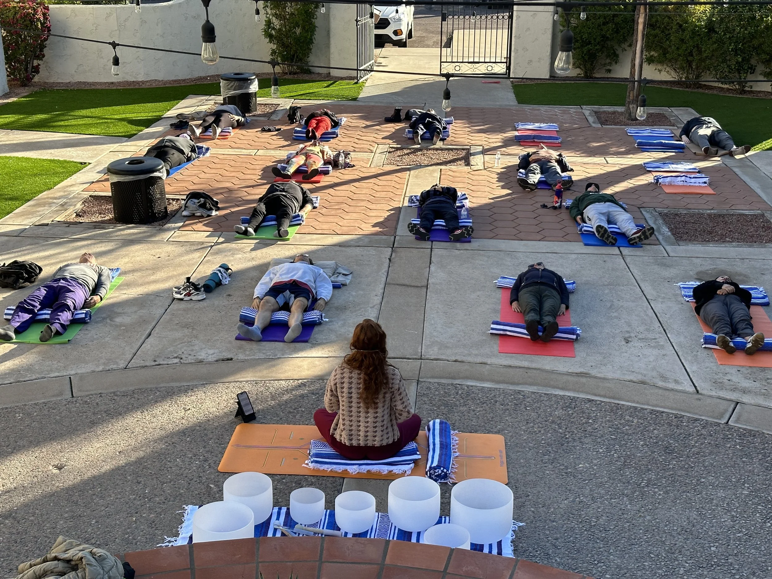 People are lying on yoga mats outdoors in a semicircle, practicing meditation or relaxation. The setting is a paved courtyard with string lights overhead, a trash bin, and some greenery and bushes along the perimeter.