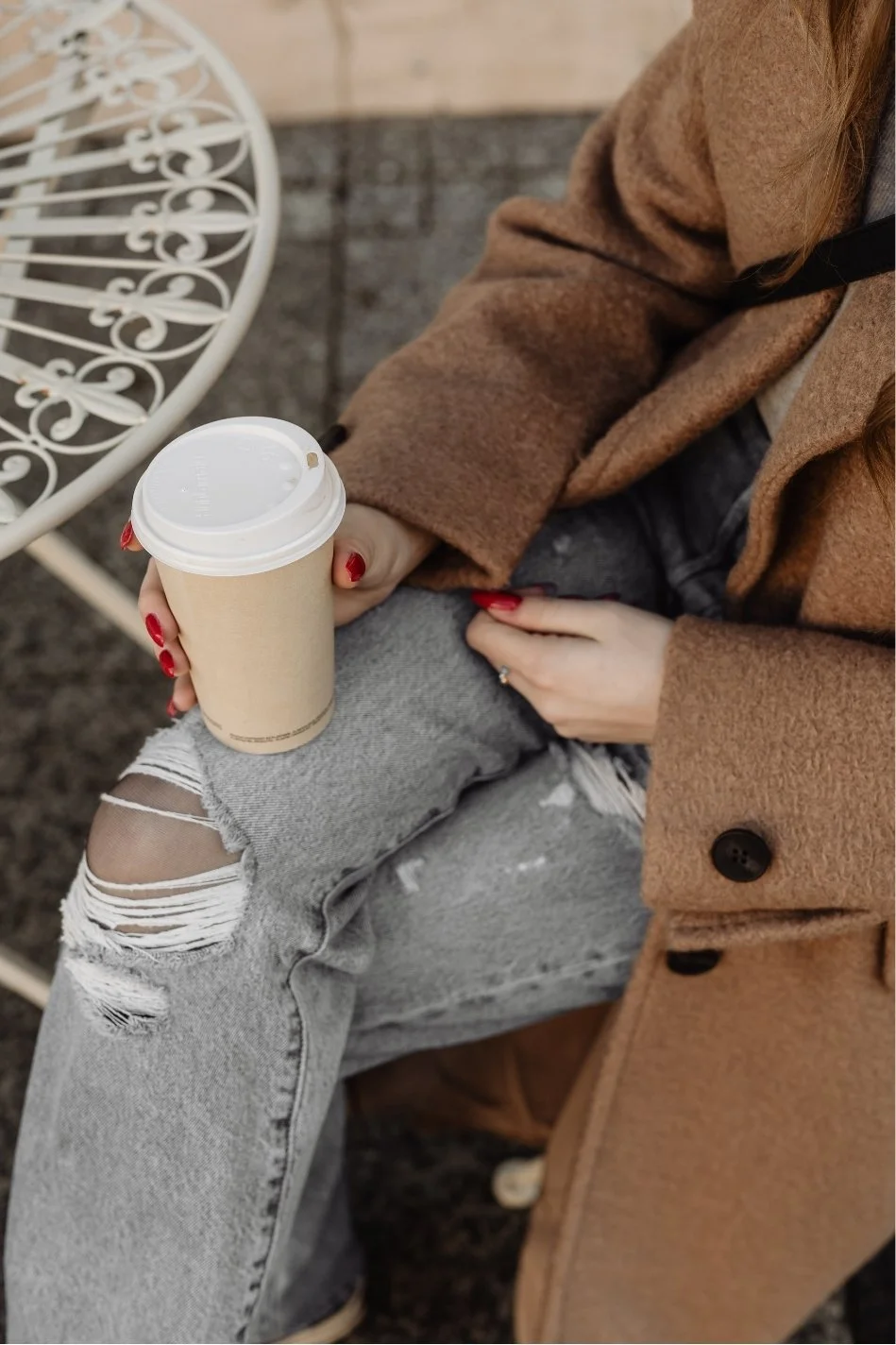 A person sitting outdoors holds a disposable coffee cup in their right hand, with red painted nails. They are wearing a brown coat, ripped light gray jeans, and have long hair.