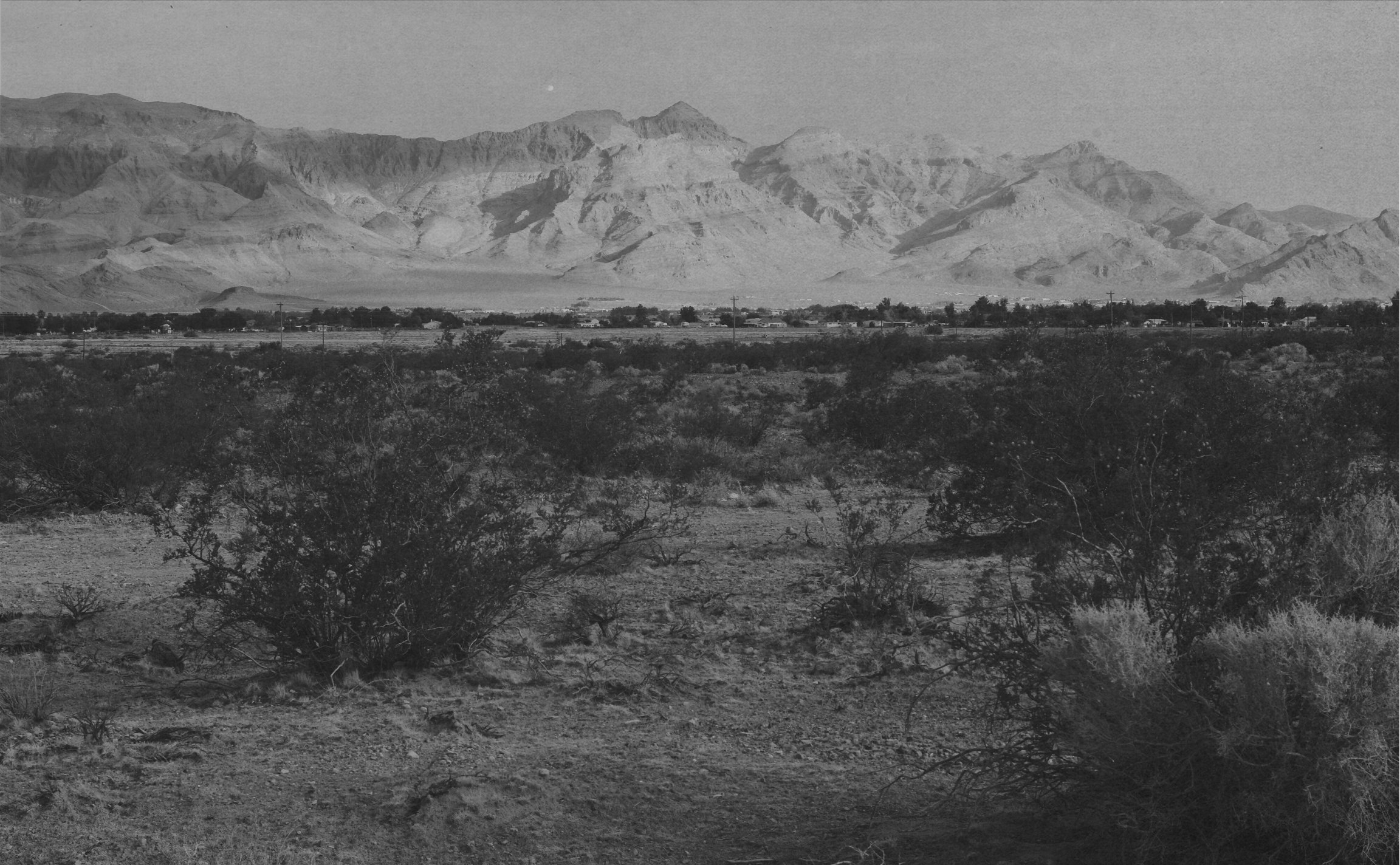 A black and white landscape photo of desert terrain with sparse bushes in the foreground, a flat area with some buildings and power lines in the middle ground, and large mountain ranges in the background.