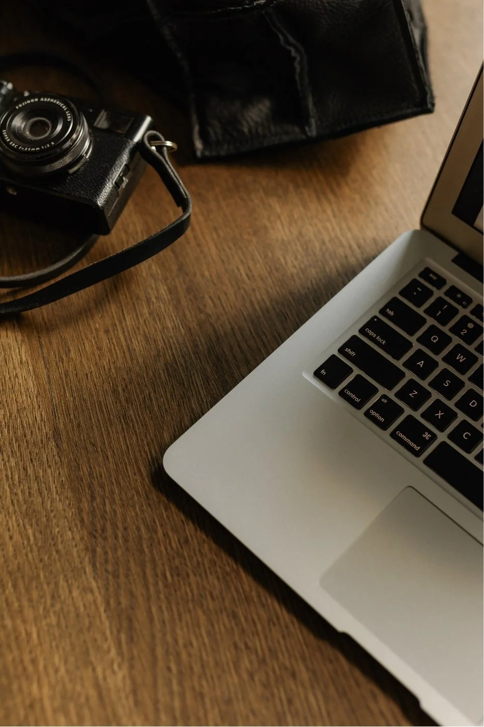 Partial view of a MacBook laptop, a compact camera with a strap, and a black leather wallet or pouch on a wooden surface.