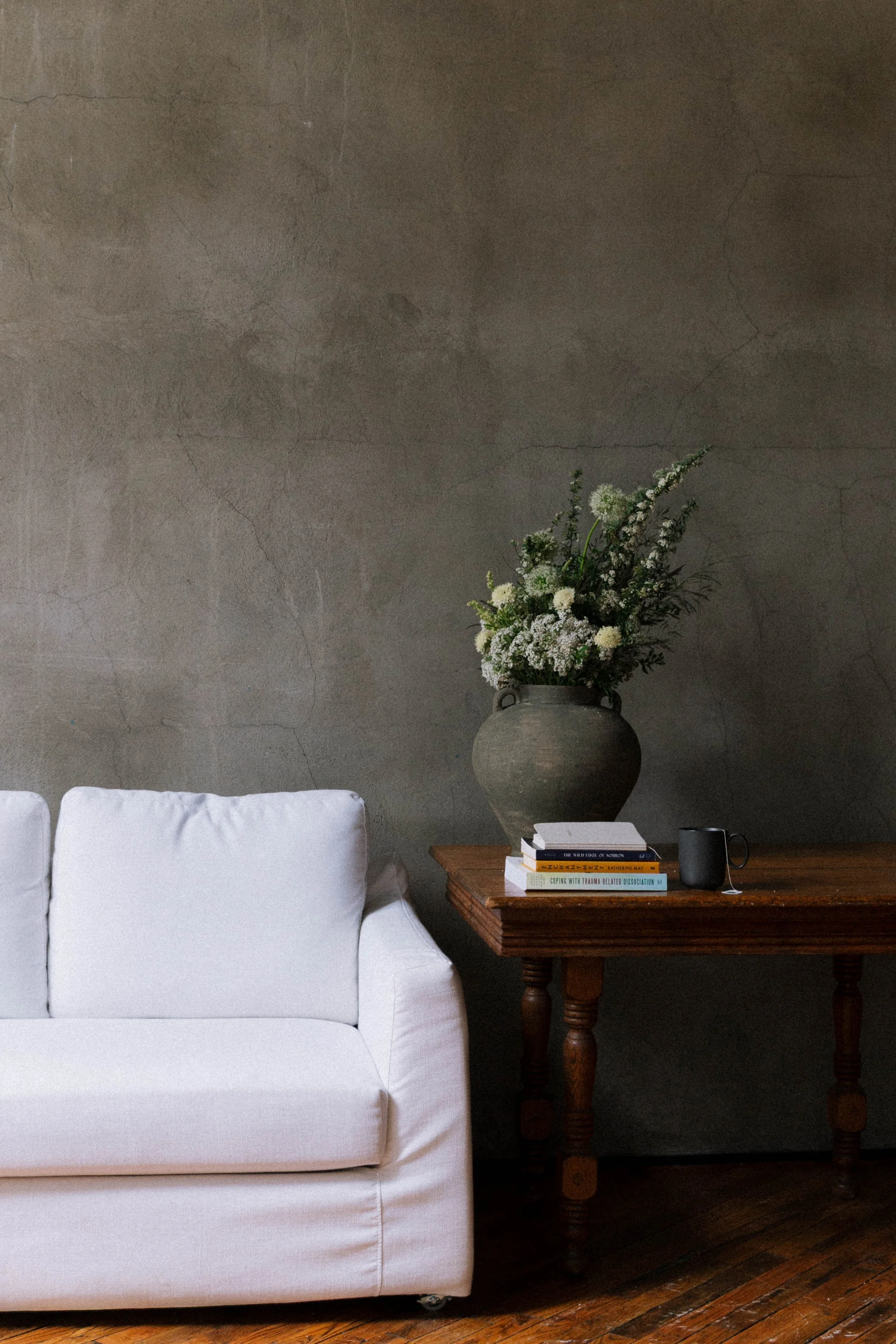 Living room scene with a white sofa, a wooden side table with books, a black mug, and a large grey vase with white and green flowers against a textured grey wall.