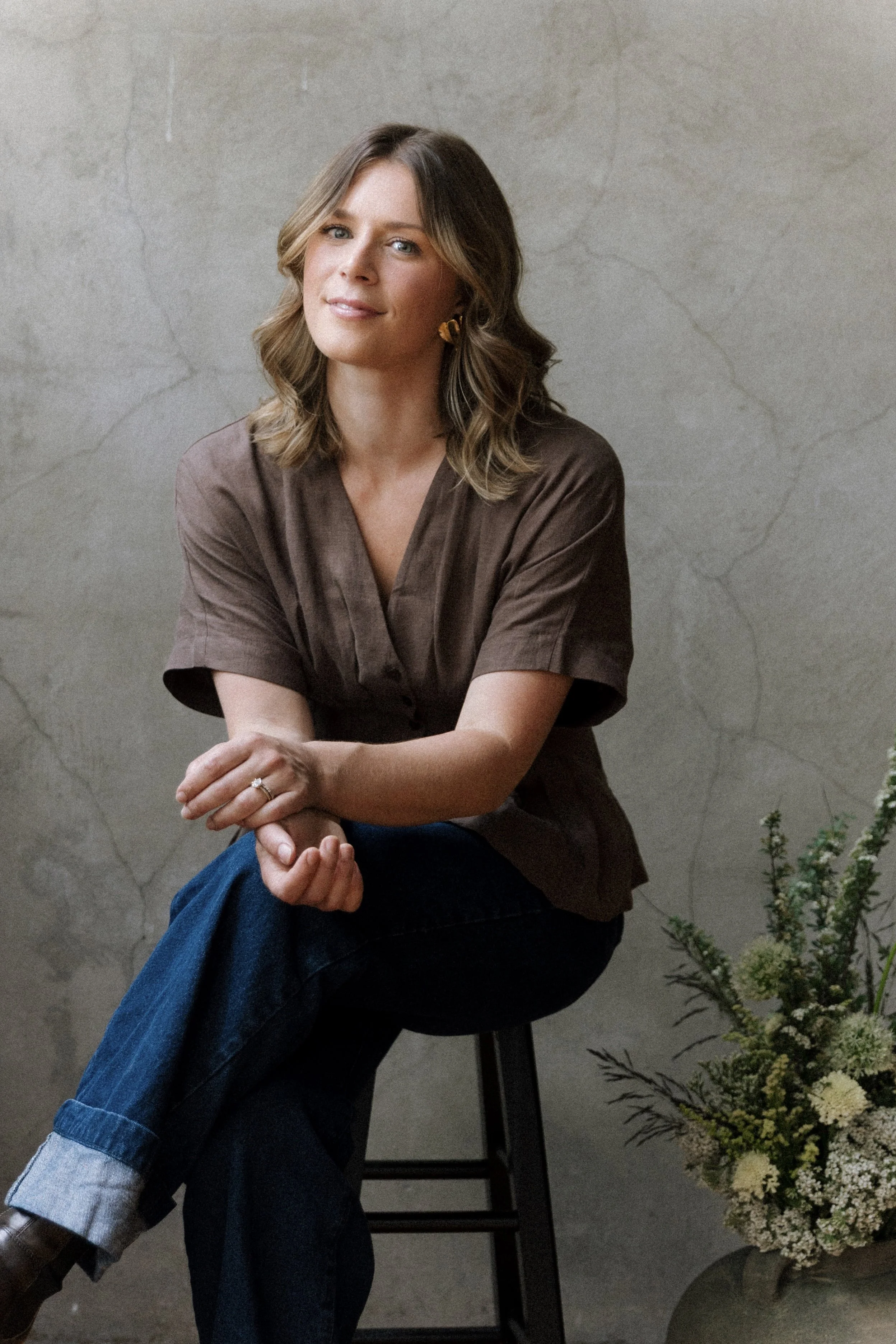 A woman with shoulder-length wavy brown hair, wearing a brown blouse and earrings, is sitting on a black stool in front of a textured beige wall. She is smiling softly with her arms crossed, and a large bouquet of yellow and white flowers is on the floor next to her.