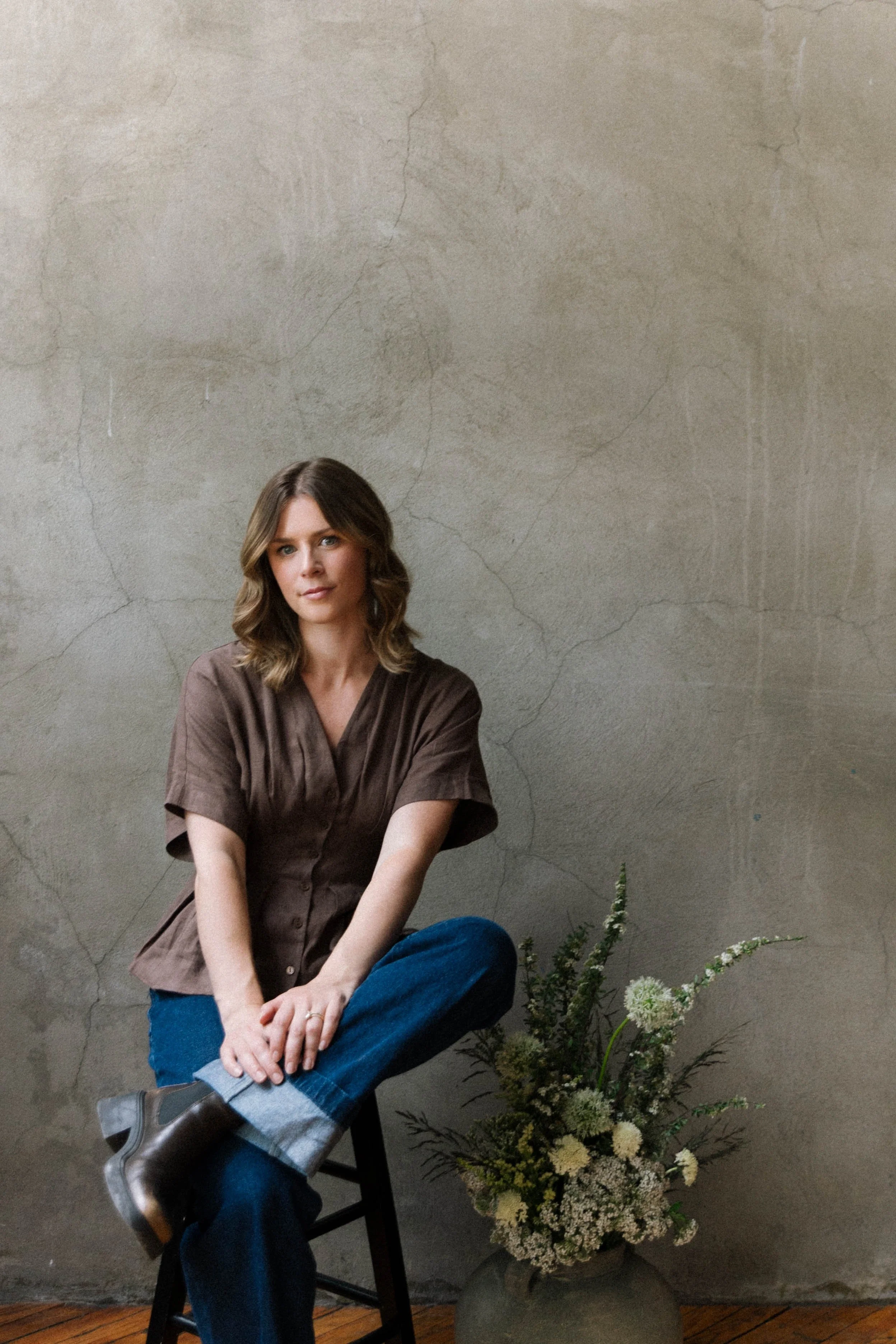A young woman with brown hair styled in loose waves, wearing a brown blouse, blue jeans, and gray boots, sits on a black chair in front of a plain gray, slightly cracked wall. She has a neutral expression and is sitting with her legs crossed and arms resting on her lap. There is a large potted plant with white flowers and greenery on the floor beside her.