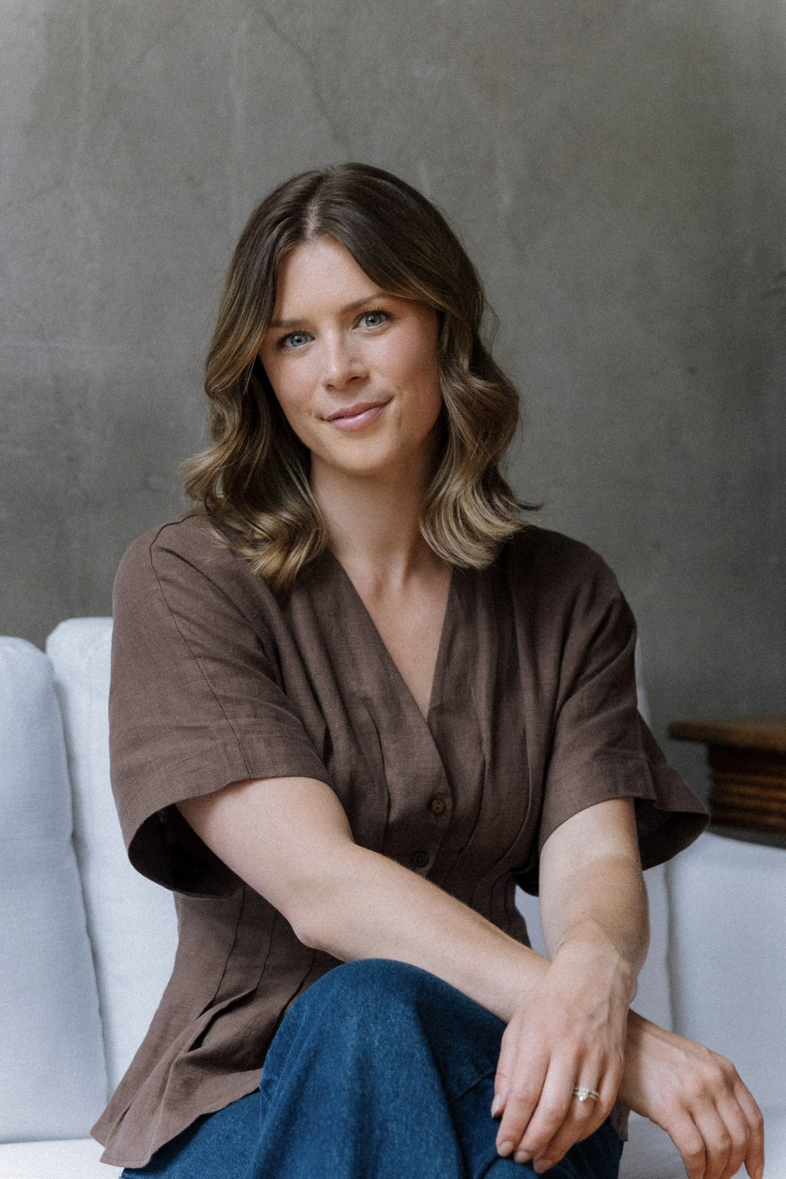 A woman with shoulder-length brown wavy hair and blue eyes sitting on a white couch, wearing a brown short-sleeved blouse and blue jeans, with a gray wall background.
