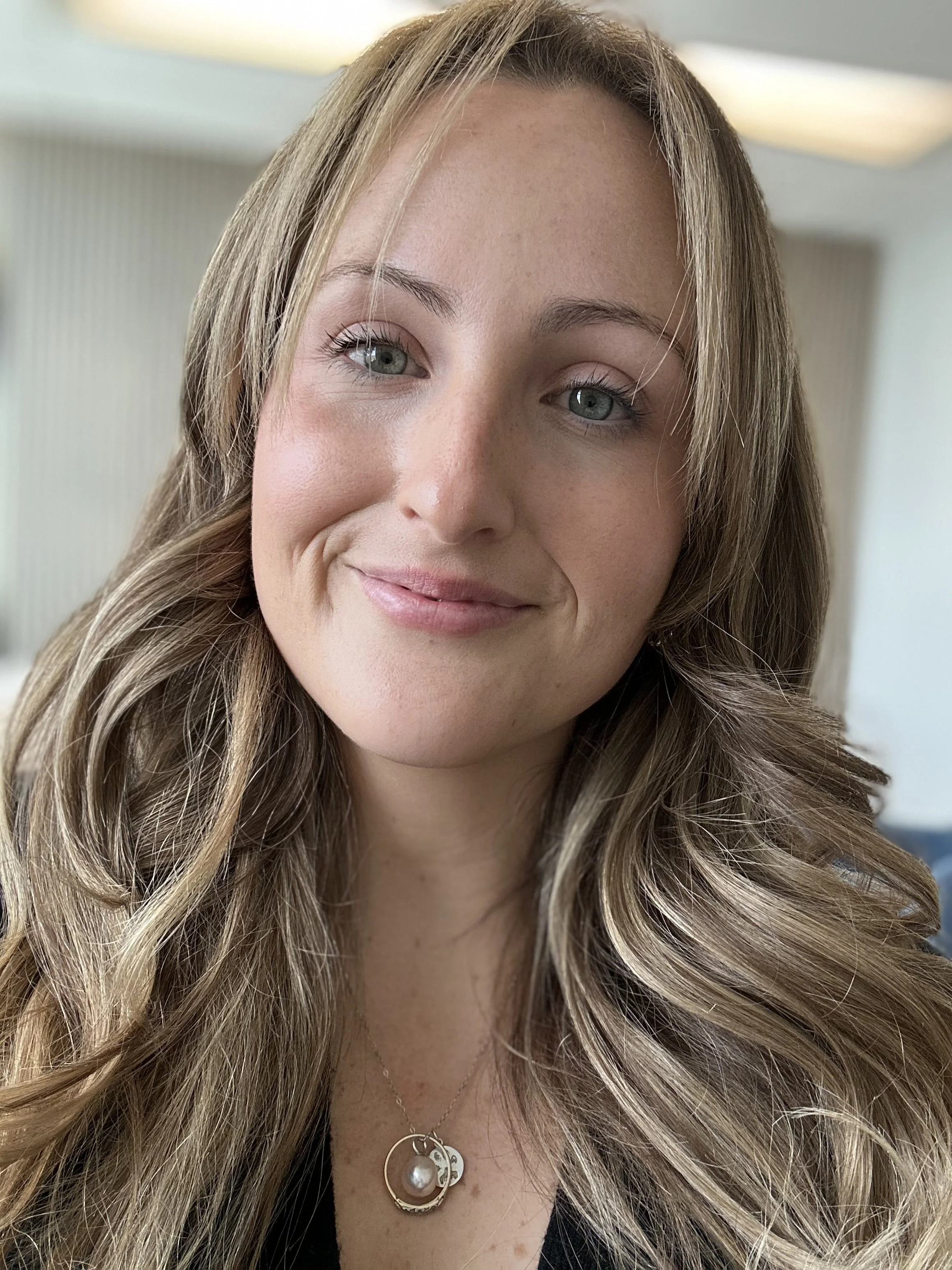 Close-up of a smiling woman with long, wavy blonde hair, wearing a gold necklace with a pearl pendant, and a black top, in an indoor setting.