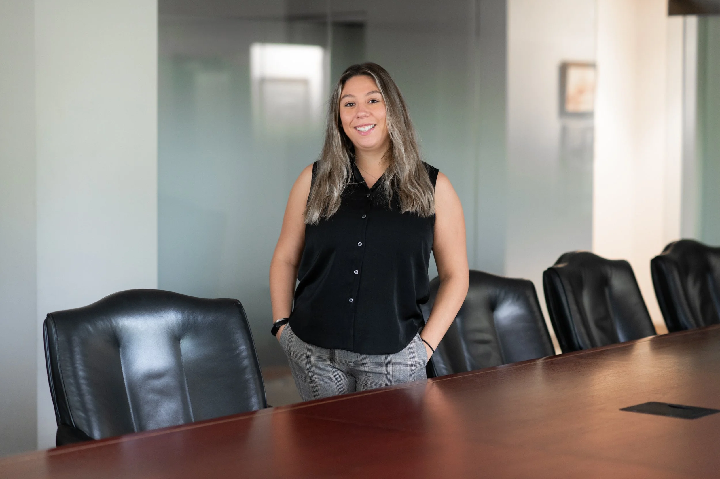 A young woman with long, wavy blonde hair wearing a sleeveless black blouse and plaid pants, smiling and standing in a conference room with black leather chairs around a large wooden table.