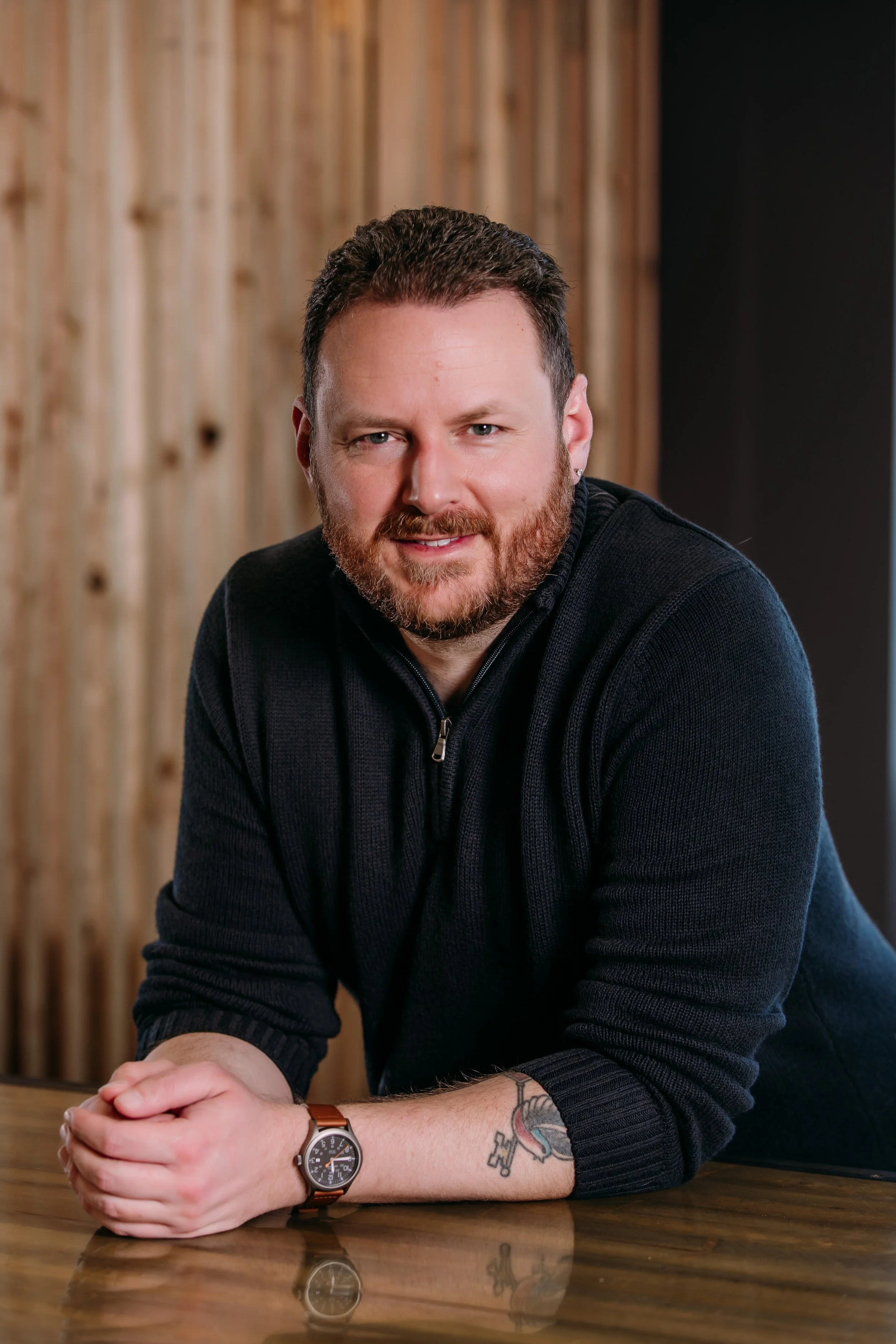 A man with a beard and tattoos on his forearm, wearing a black sweater and a watch, leaning on a wooden table with his hands clasped together, in front of a wooden wall.