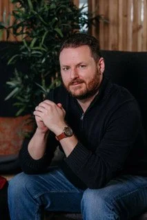 A man with a beard and short hair, wearing a black sweater and blue jeans, sitting indoors with his hands clasped, in front of a wooden wall and green plants.