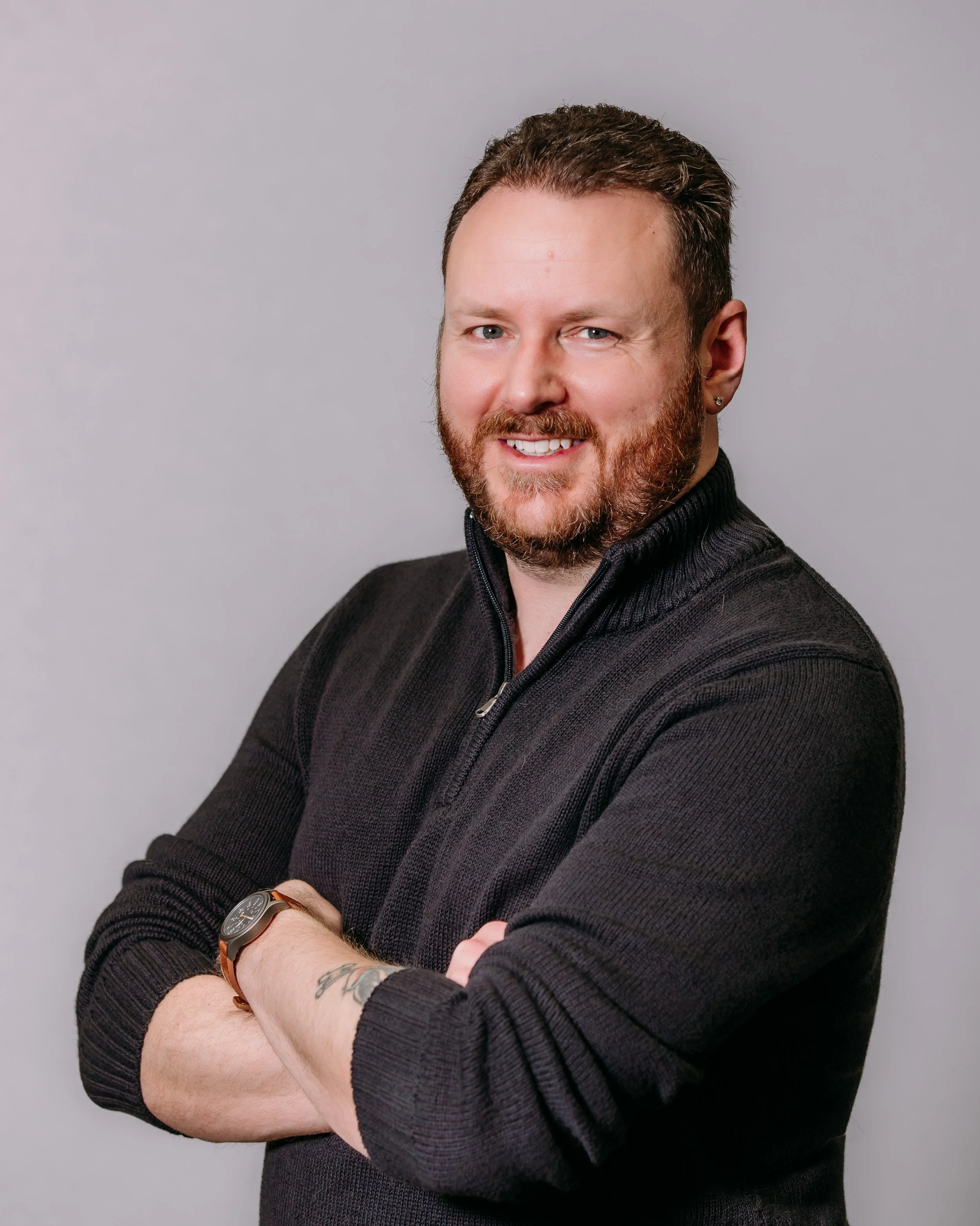 Jonathan P. Morrow smiling with arms crossed, wearing a black zip-up sweater and a watch, against a plain gray background.