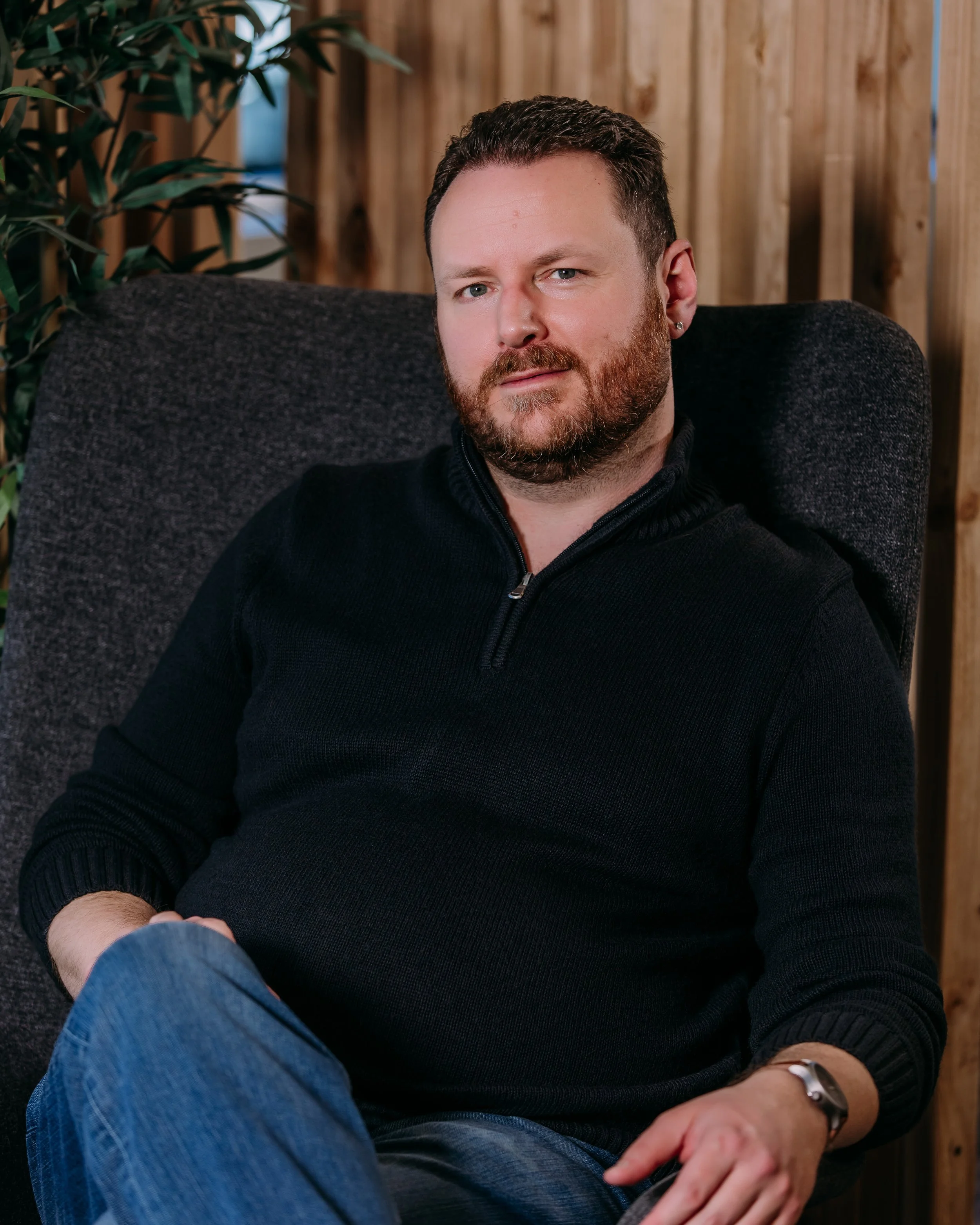 A man with a beard and earrings sitting on a dark gray chair with a wood-paneled wall and a plant in the background.