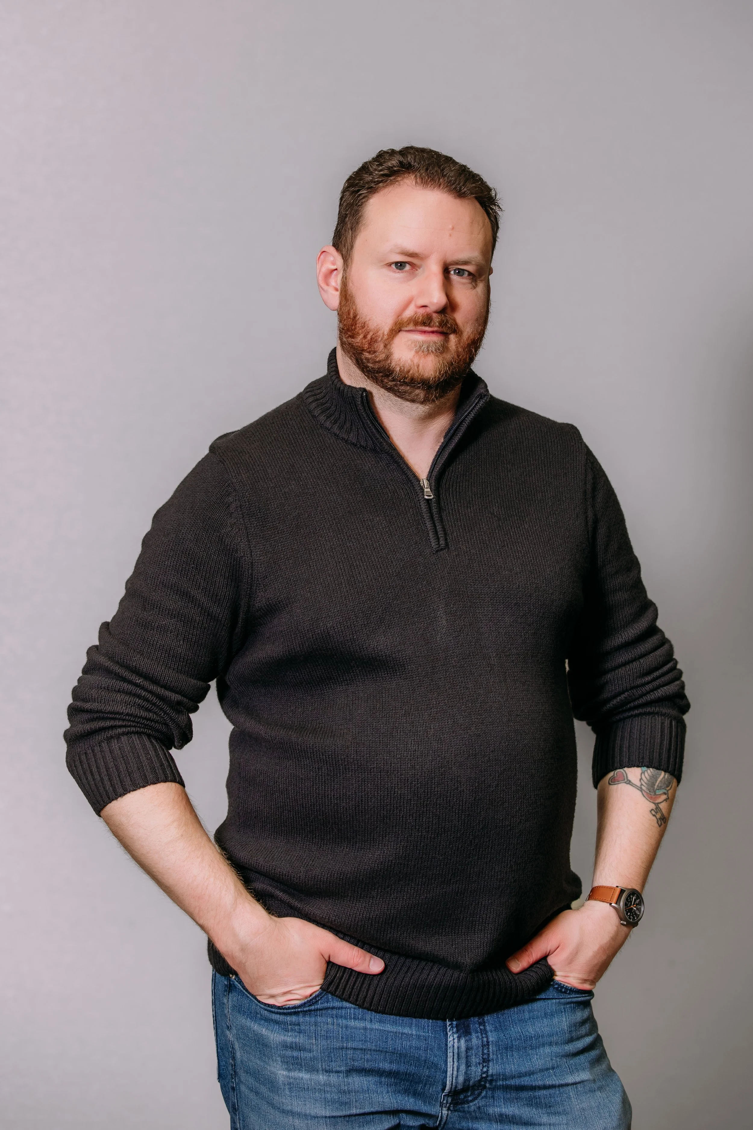 A man with a beard and short hair, wearing a black zip-up sweater with the zipper partially open, blue jeans, and a wristwatch, standing against a plain light gray background with his hands in his pockets.